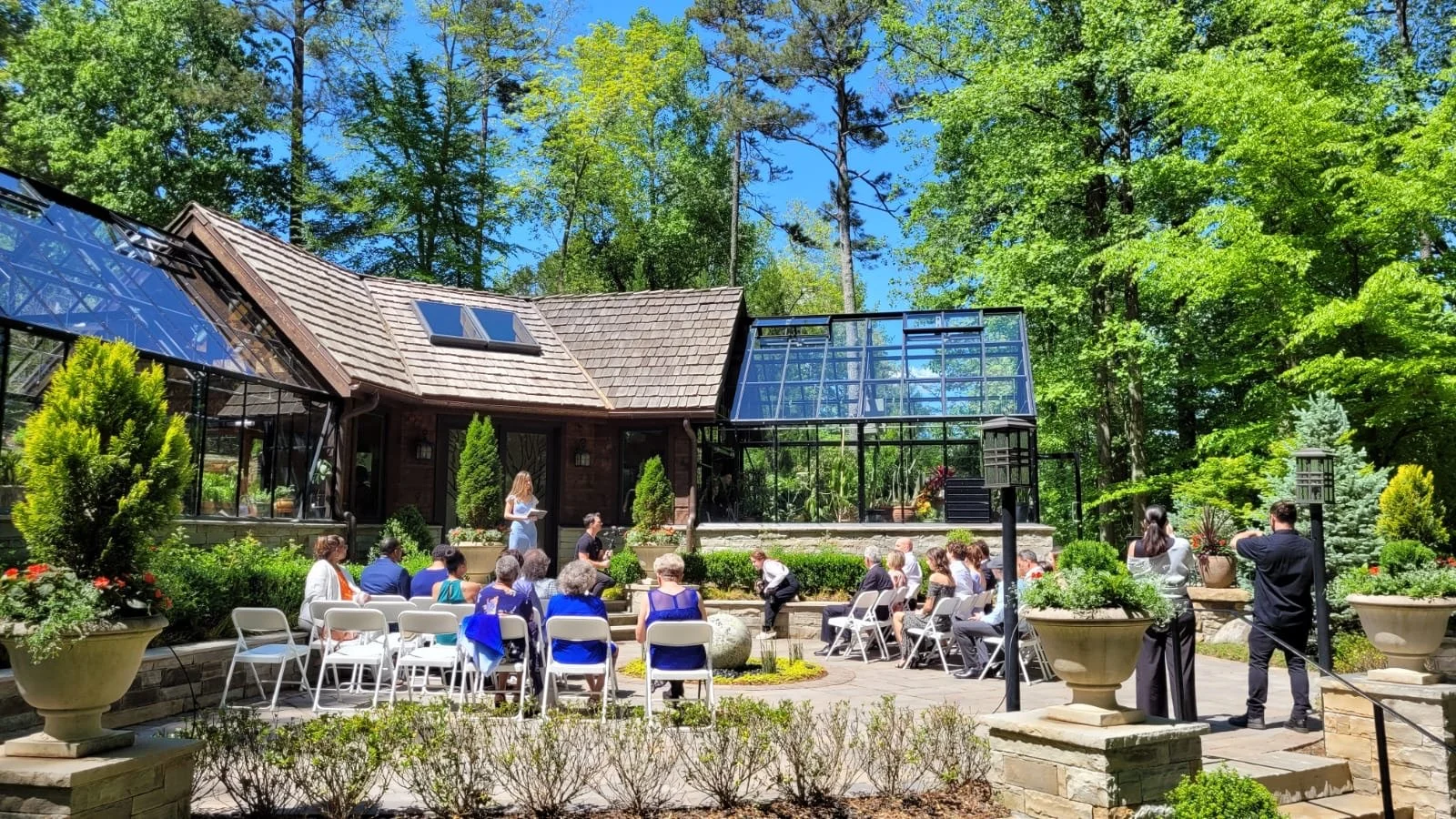 Guests attending an outdoor wedding ceremony in a lush garden with a house and glass greenhouse in the background on a bright, sunny day.