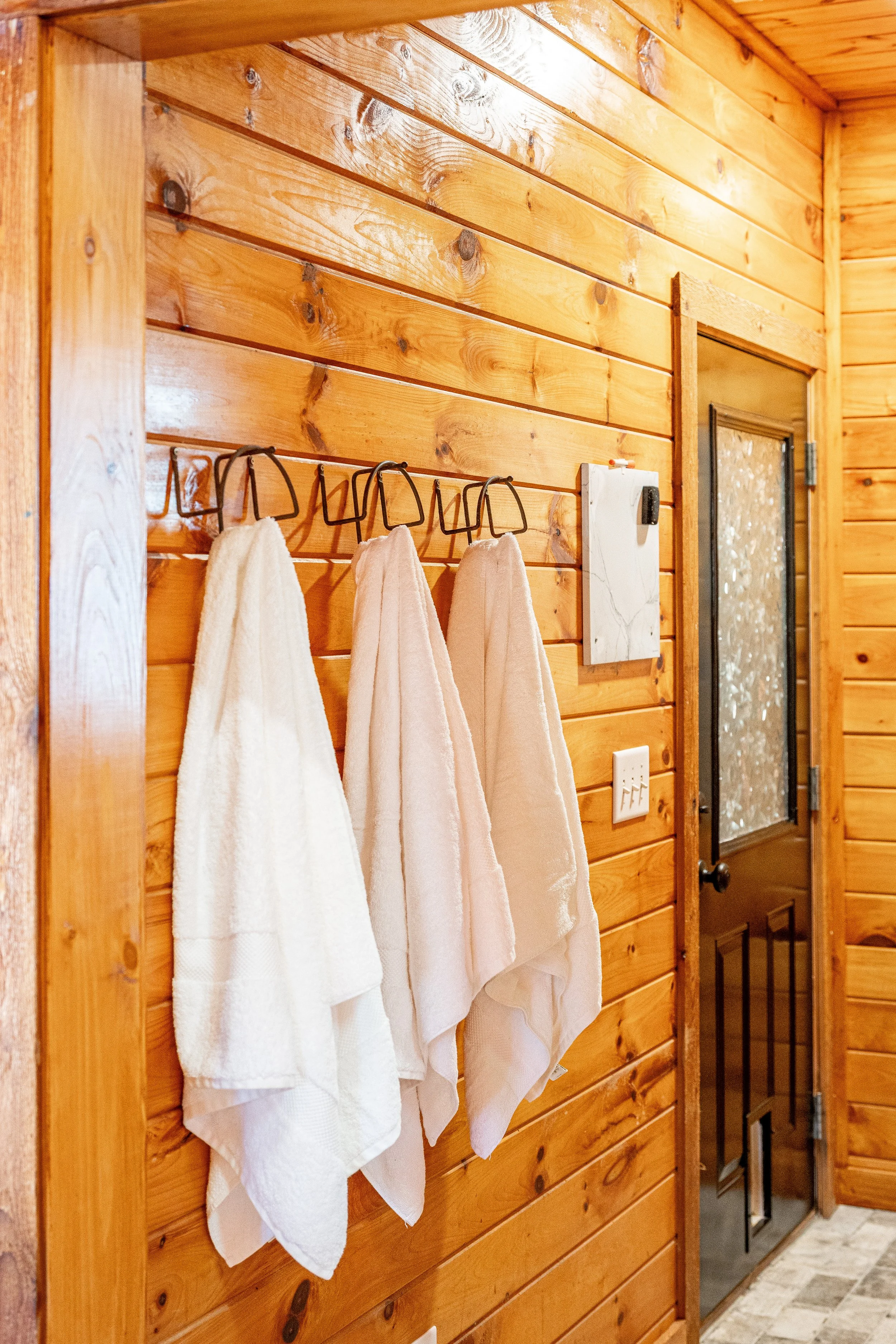 Three white towels hanging on hooks on a wooden wall in a bathroom near a door with a glass panel.