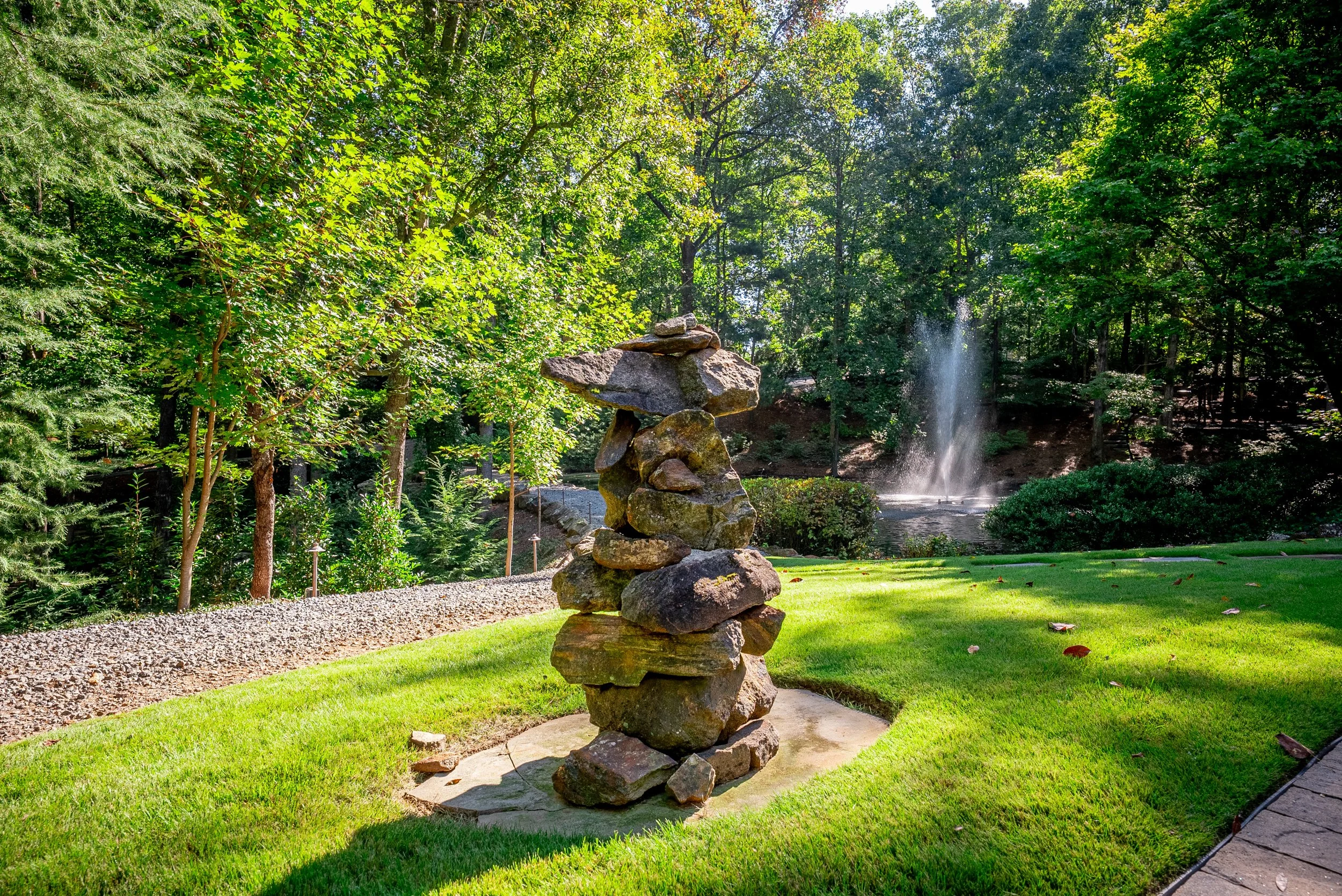 Stacked rocks in a garden with a fountain and lush green trees in the background.