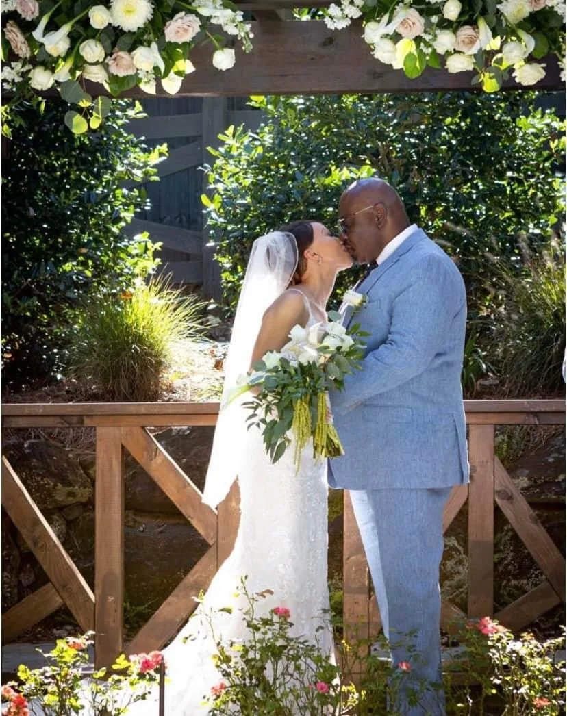 A bride and groom kissing outdoors beneath a floral arch at their wedding, surrounded by greenery and colorful flowers.