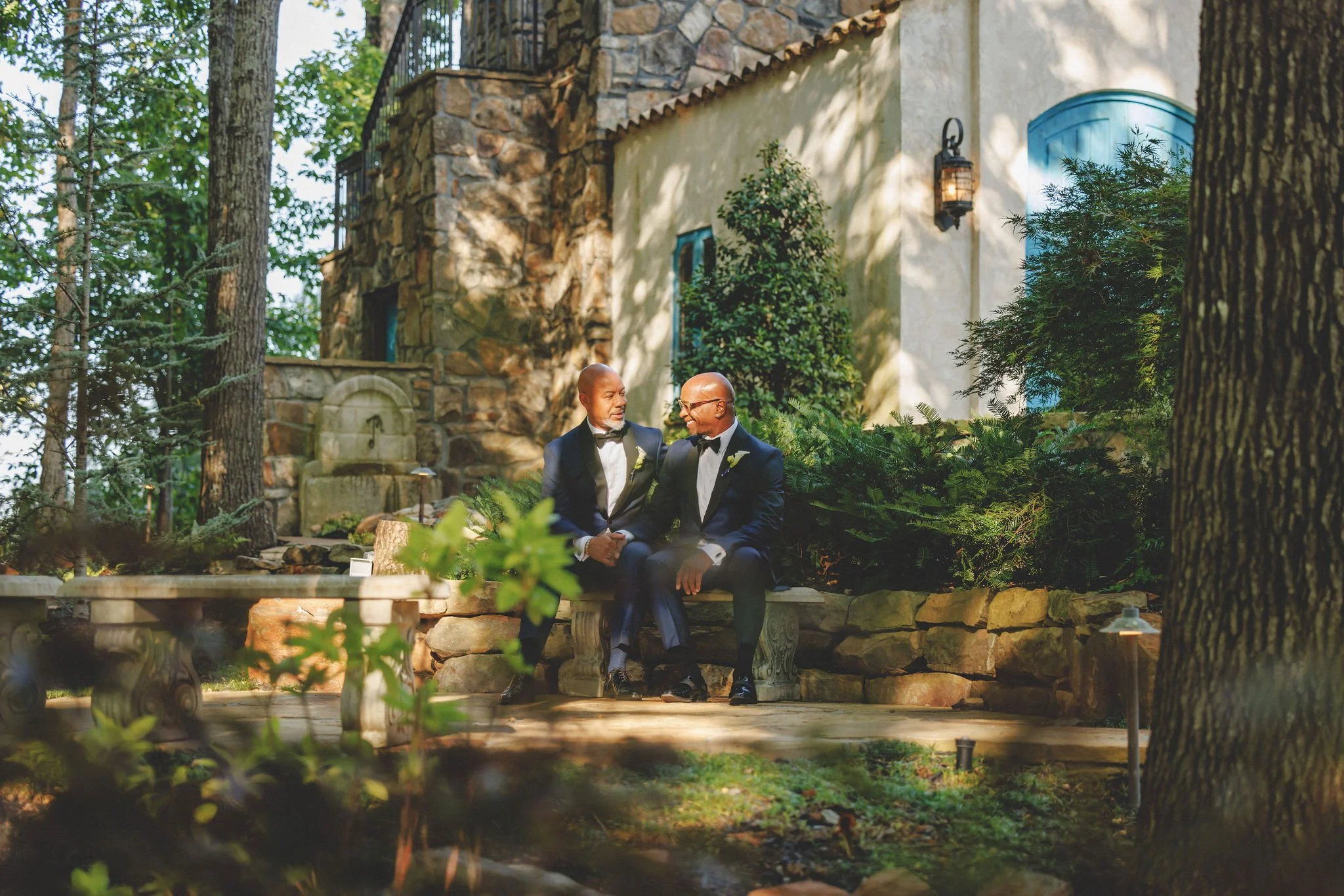 Two men dressed in black tuxedos, sitting on a stone bench outdoors, engaging in conversation, with a stone building and trees in the background.