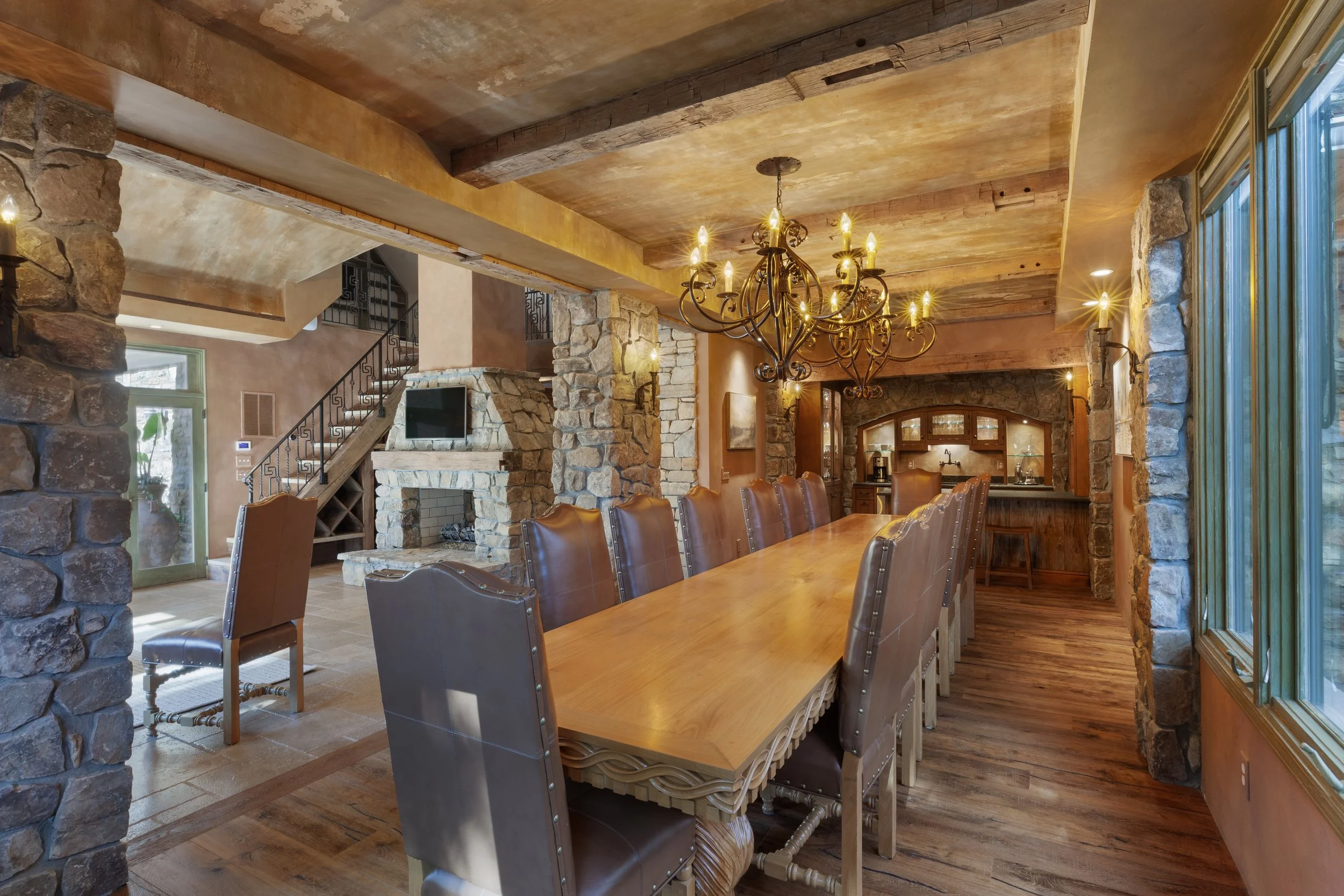 A spacious dining room with rustic stone walls, a large wooden dining table, and leather chairs. An ornate chandelier hangs above the table, and a stone fireplace is visible in the background. Large windows let in natural light.