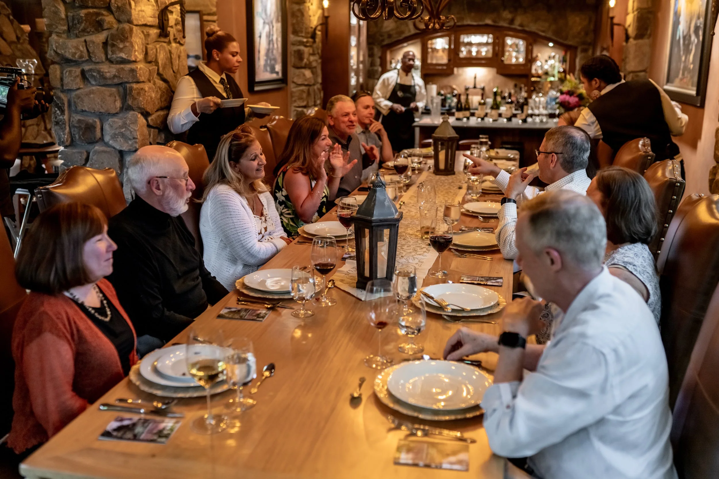 People gathered around a long dining table in a restaurant or private dining room, with waitstaff serving in the background.