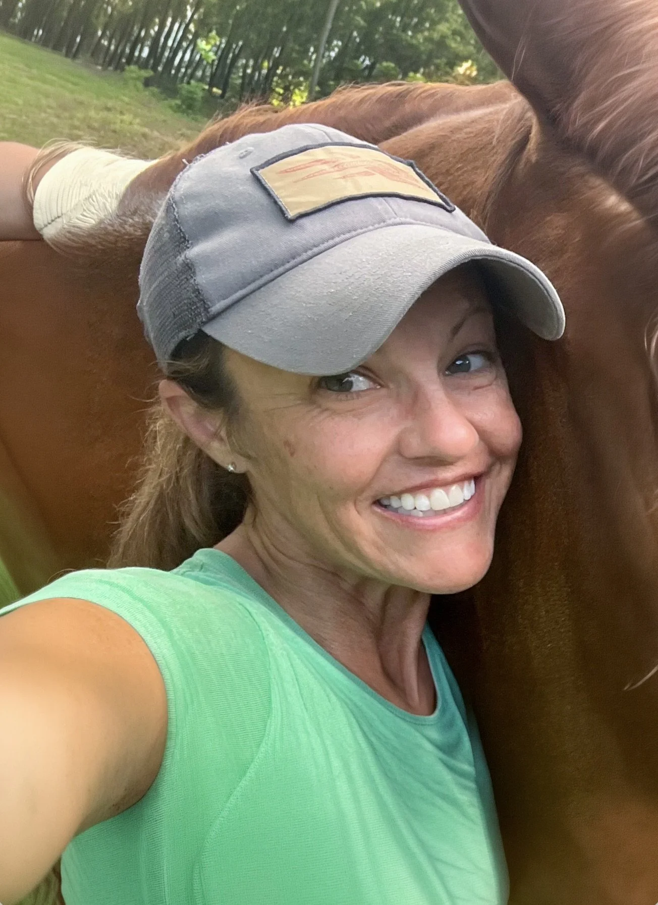 A woman smiling with a horse in a green outdoor area, wearing a gray cap and a light green shirt.