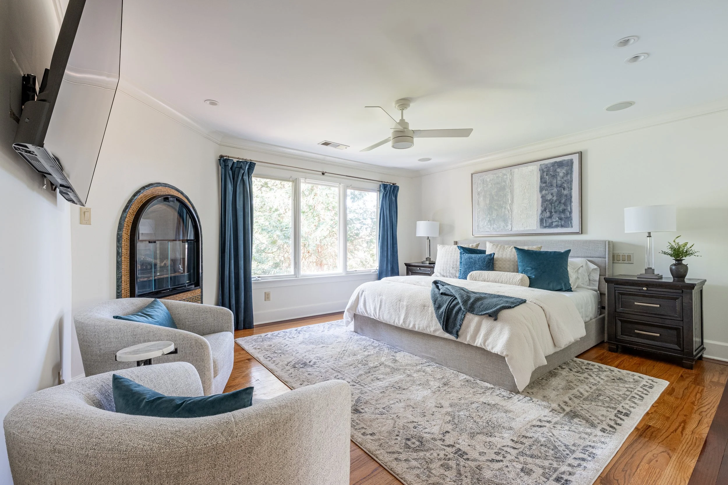 A spacious bedroom with a large bed, blue pillows, and a gray headboard, flanked by two dark nightstands with lamps. A window with blue curtains lets in natural light, and there's a comfortable armchair near a fireplace. A modern artwork hangs above 