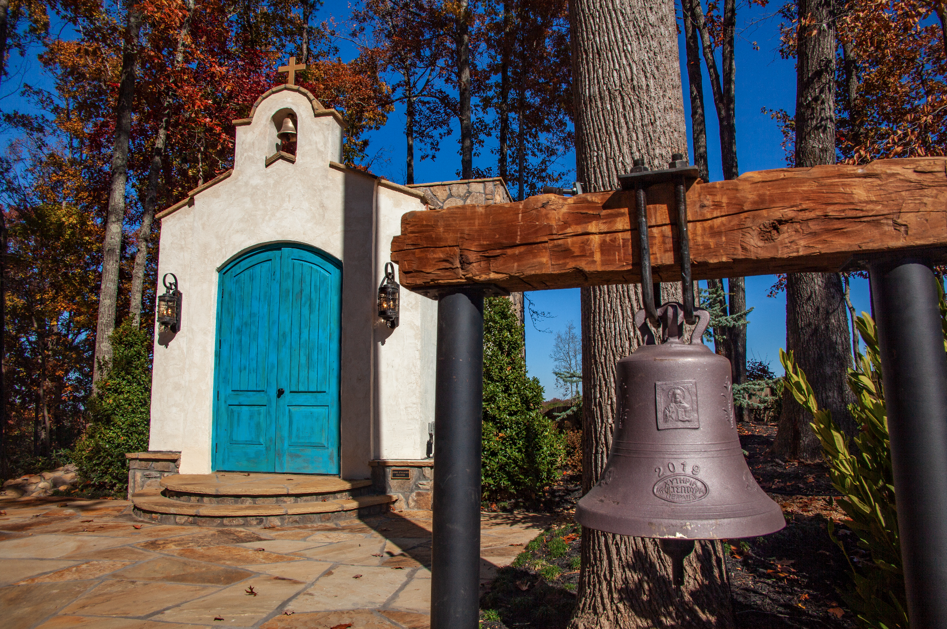 Small white chapel with a bright blue wooden door, iron lanterns on either side, a small bell tower with a bell and cross, and an old-fashioned bronze bell hanging from a wooden beam, set in a wooded area with tall trees and a clear blue sky.