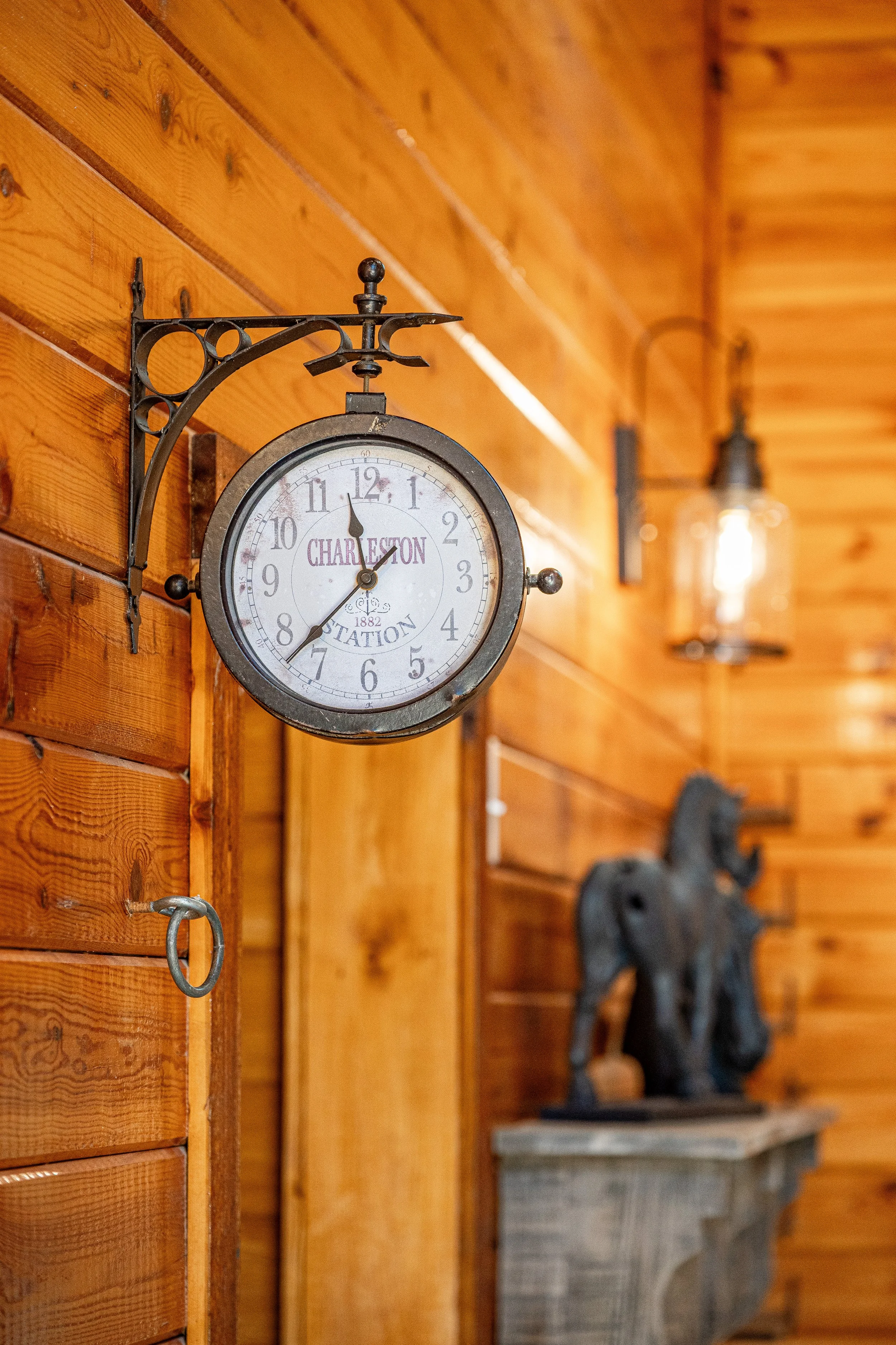 A vintage station clock mounted on a wooden wall, showing the time as 11:57. The clock has the words 'Charleston Station' and the date 1882 on its face. The background features warm-toned wooden paneling and an out-of-focus elephant sculpture on a pe