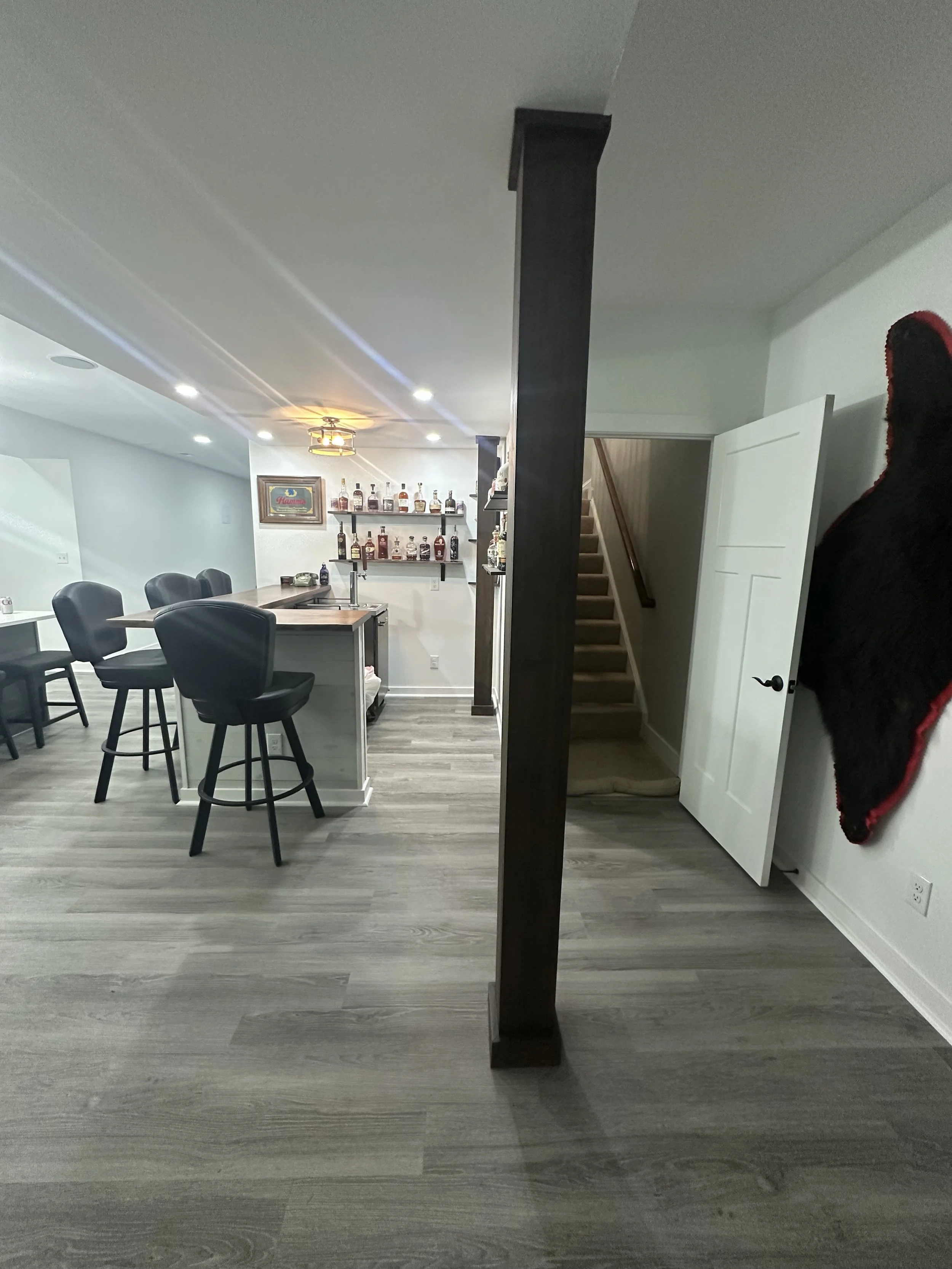 Interior view of a modern open-concept living space with a bar area featuring high chairs, a staircase leading to an upper floor, and a wall-mounted shelf with liquor bottles.