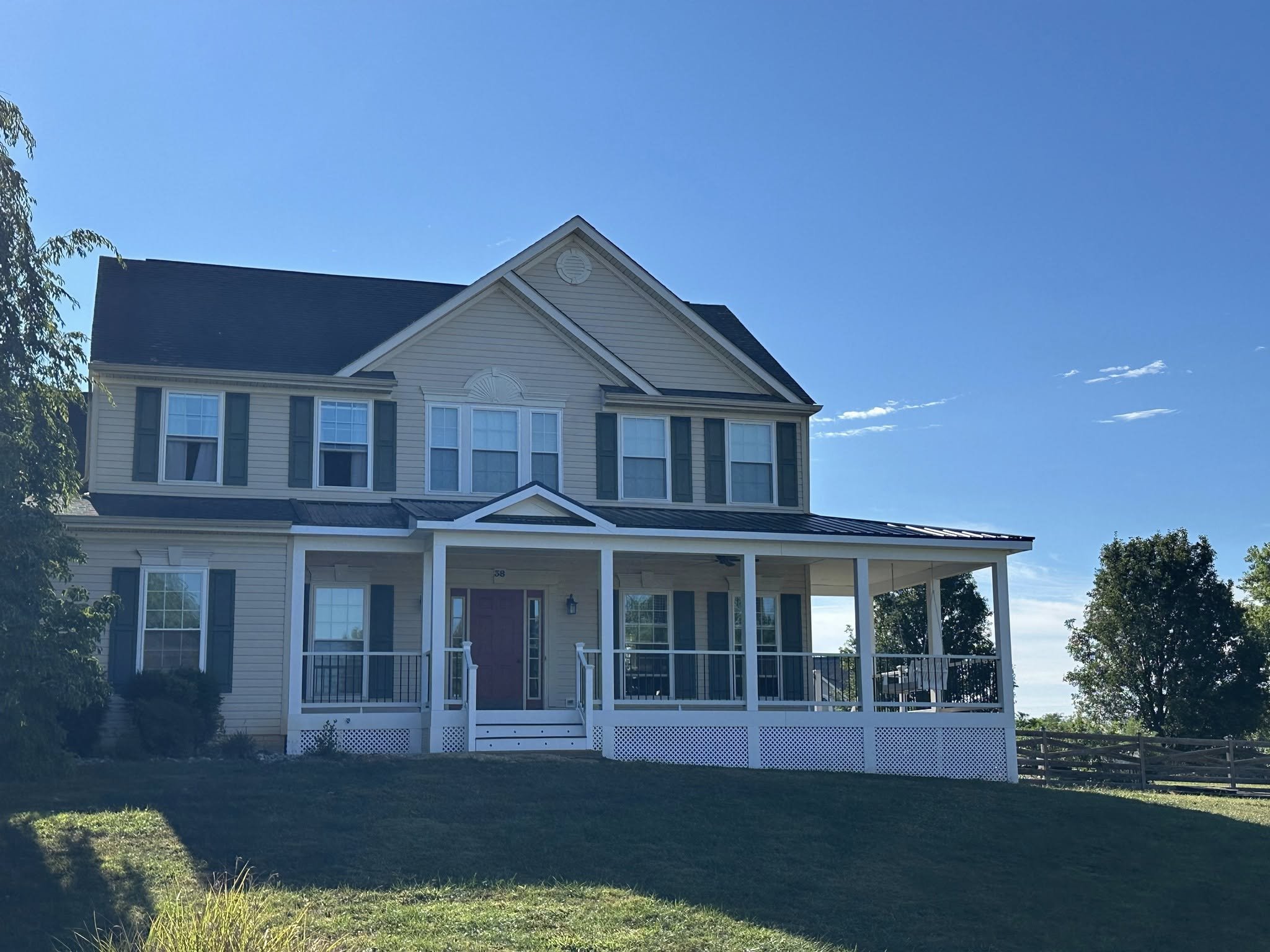 Two-story house with beige siding, white trim, a front porch with white railings, and dark green shutters. The house is set on a grassy hill with trees and a fence in the background, under a blue sky with some clouds.