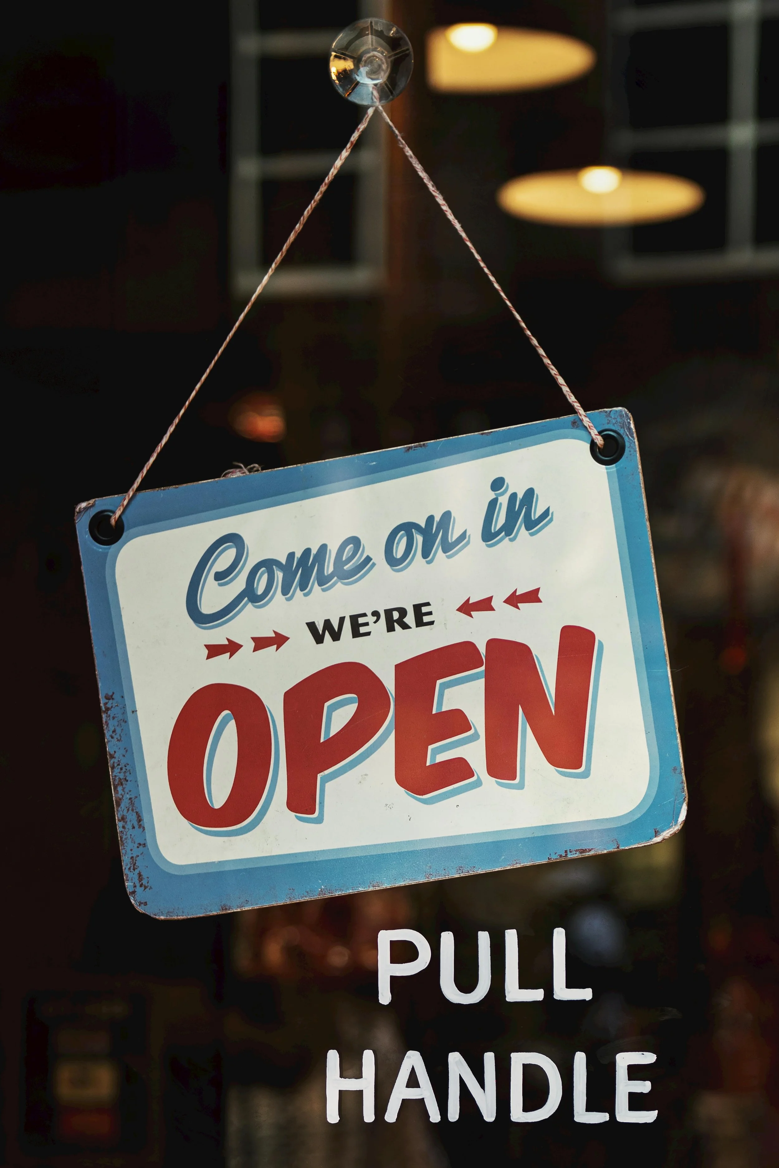 A blue and white sign hanging with a string reads 'Come on in, we're OPEN' with an 'Open' sign in large red letters, and below it, a smaller white sign says 'PULL HANDLE' in black letters. The background appears to be the interior of a store or restaurant.