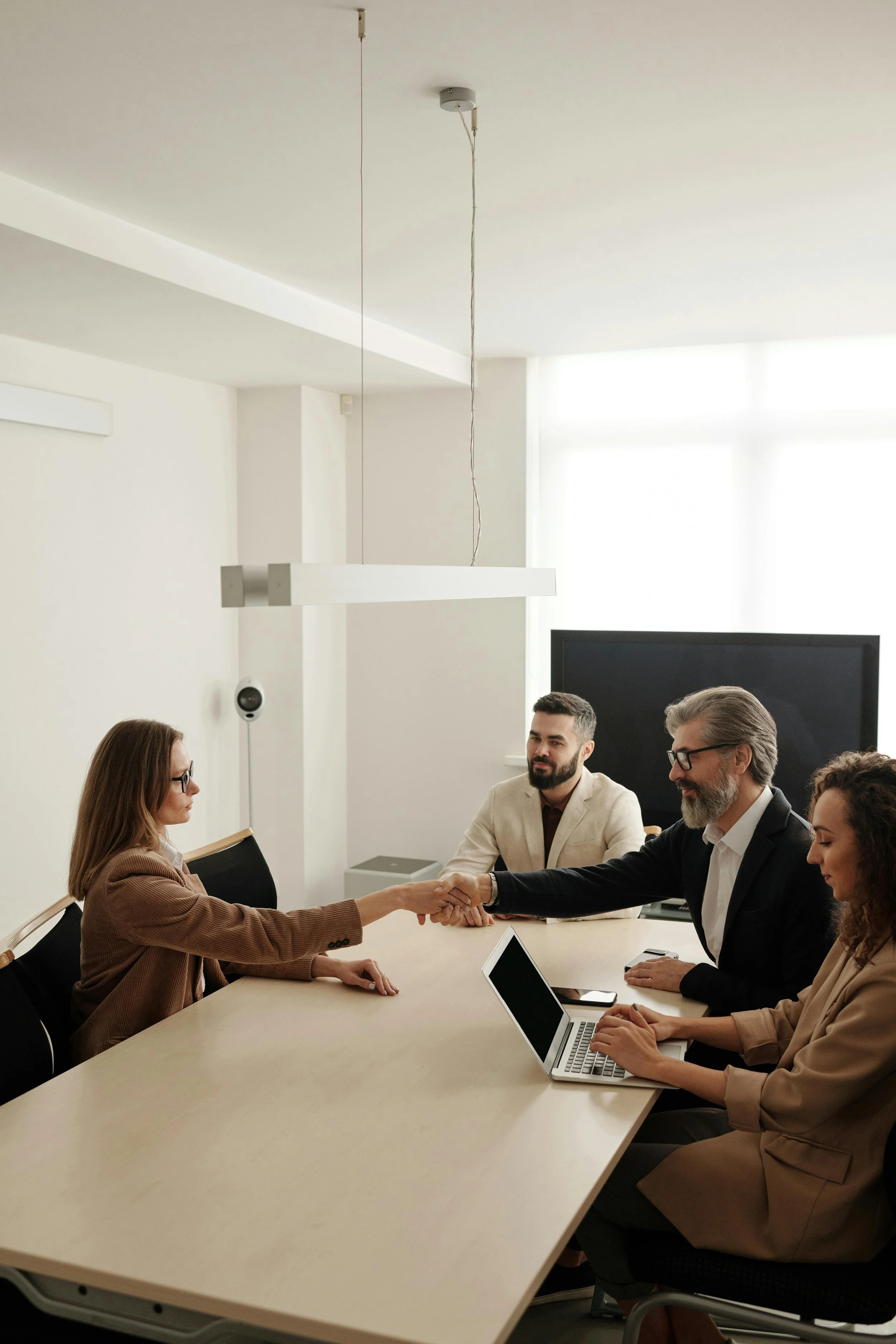 Five people in business attire sitting around a conference table, with a woman shaking hands with an older man, in a modern office with a large window and a monitor in the background.