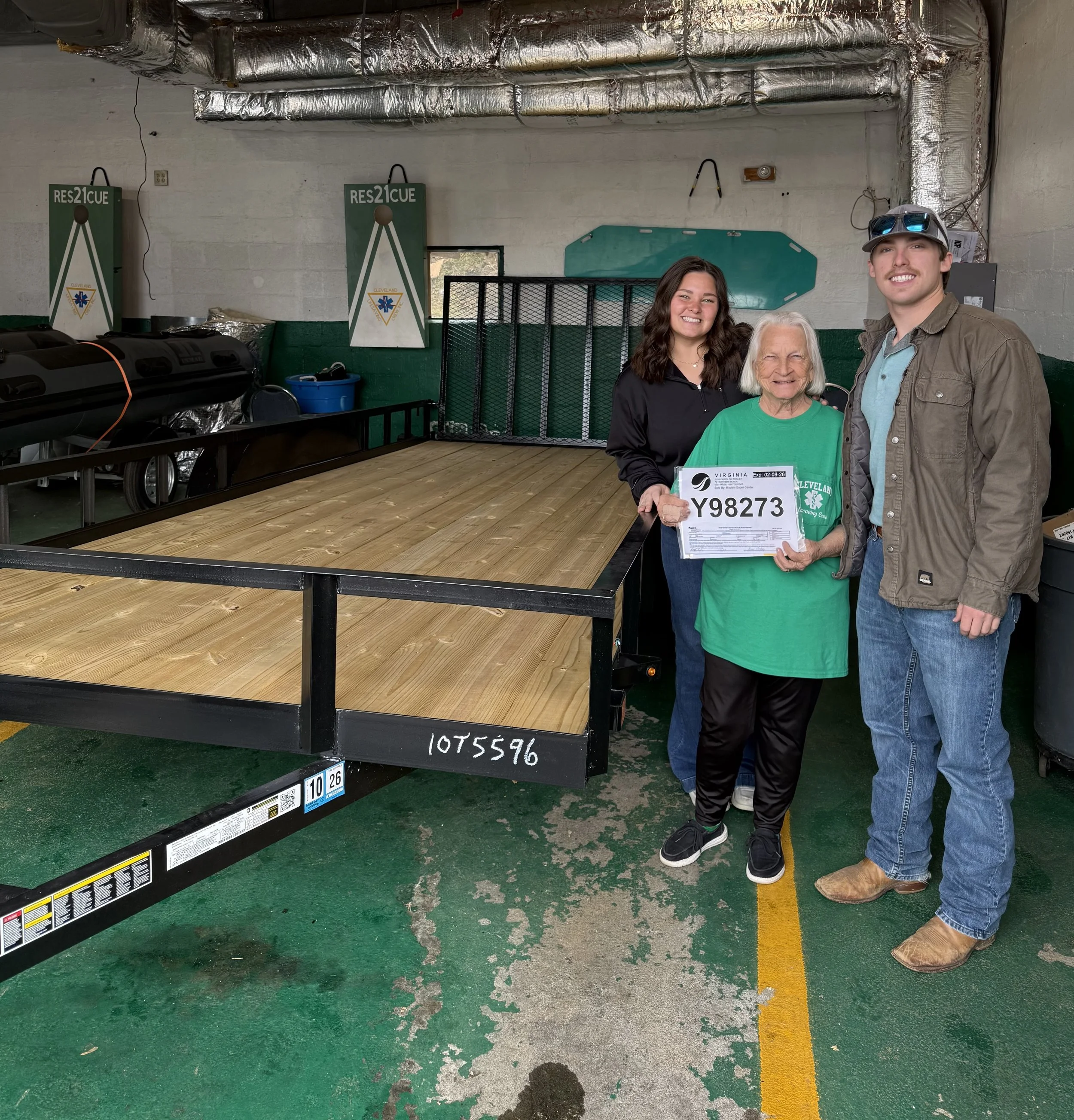 Three women standing together inside a garage, one holding a Virginia lottery scratch-off ticket and smiling. A large wooden flatbed trailer is in the background, along with garage equipment and vents.