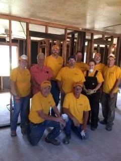 Group of ten construction workers in a partially built structure, wearing yellow shirts and safety glasses.
