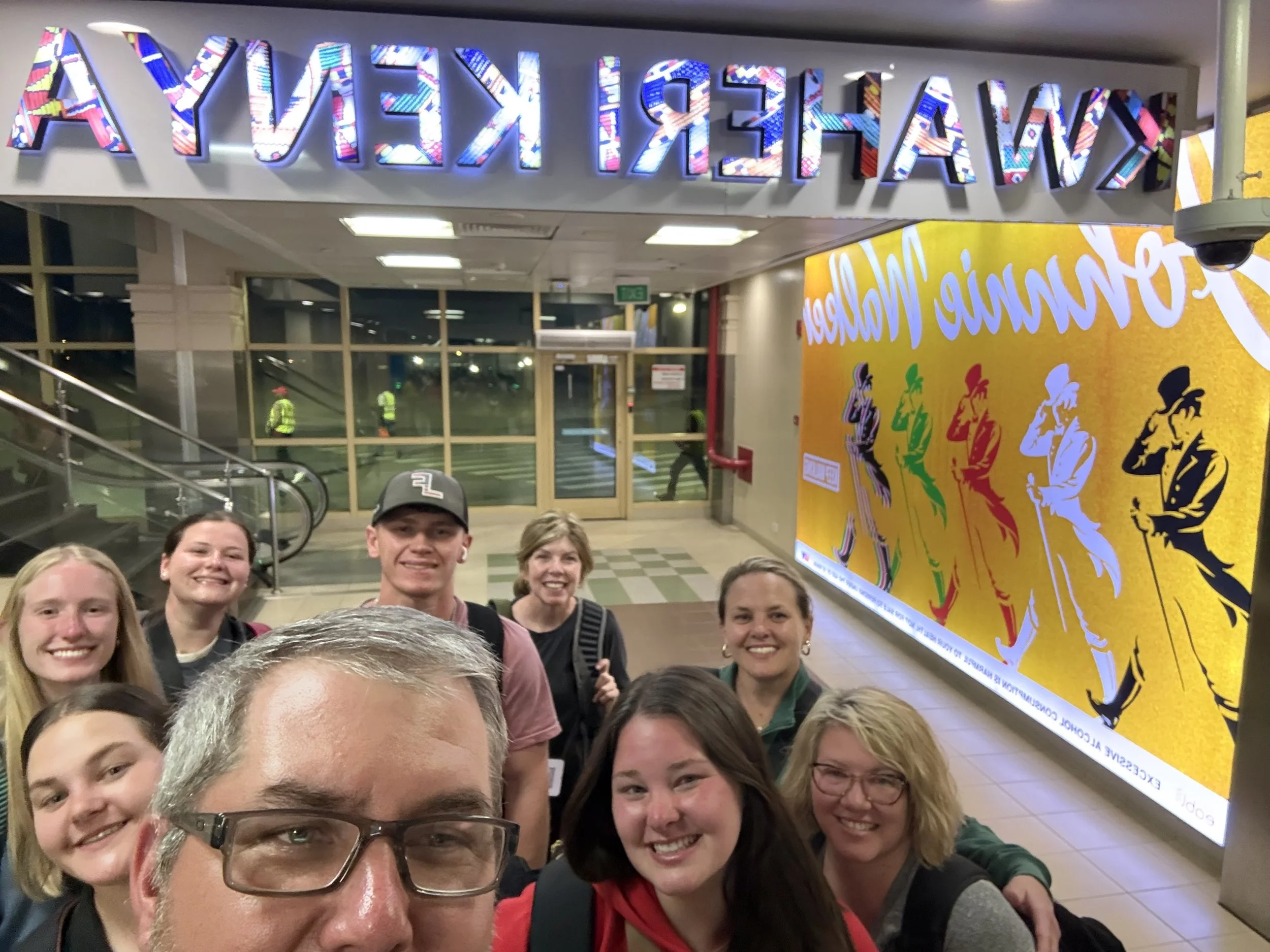 Group of people smiling for a selfie in front of a colorful display at an airport terminal.