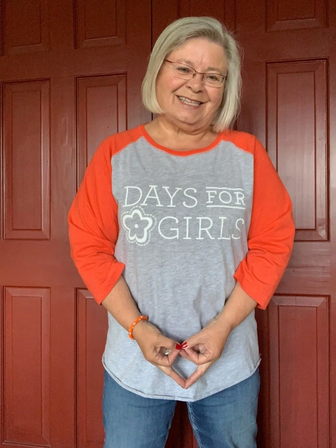 A smiling woman with short gray hair and glasses, wearing a gray and orange baseball tee that reads "DAYS FOR GIRLS," standing in front of a red wooden door.