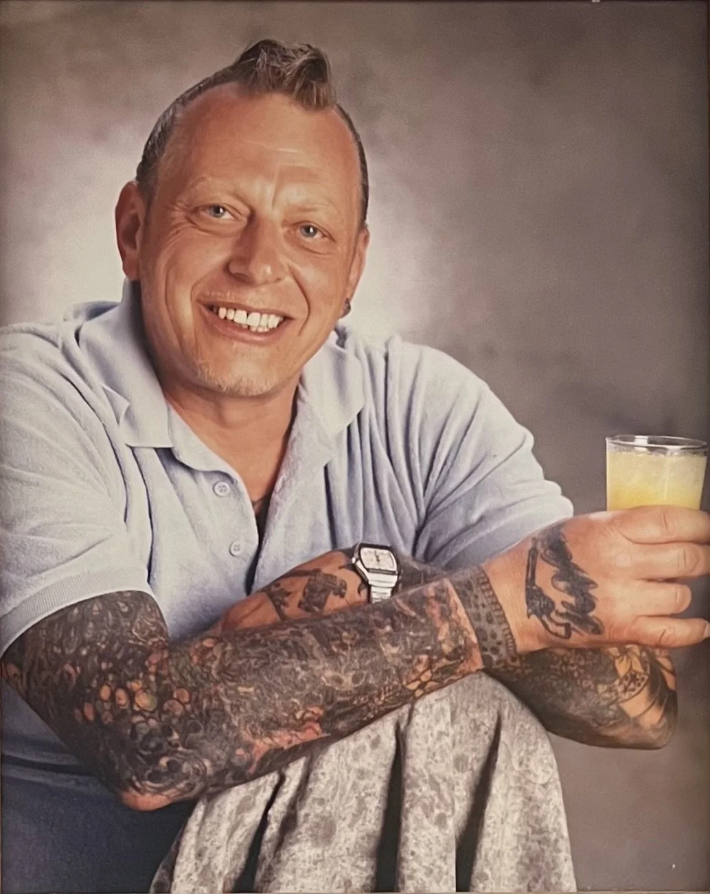 A smiling man with tattoos on his arms, wearing a light-colored polo shirt, holds a glass of drink, seated with arms crossed, against a neutral background.
