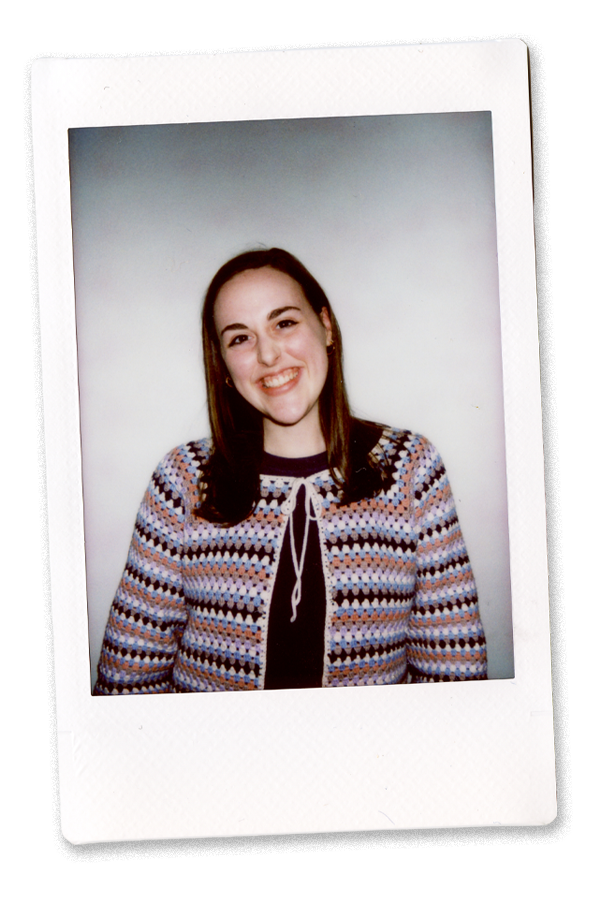 Young woman smiling with shoulder-length brown hair, wearing a colorful patterned sweater over a black top, against a plain white background.
