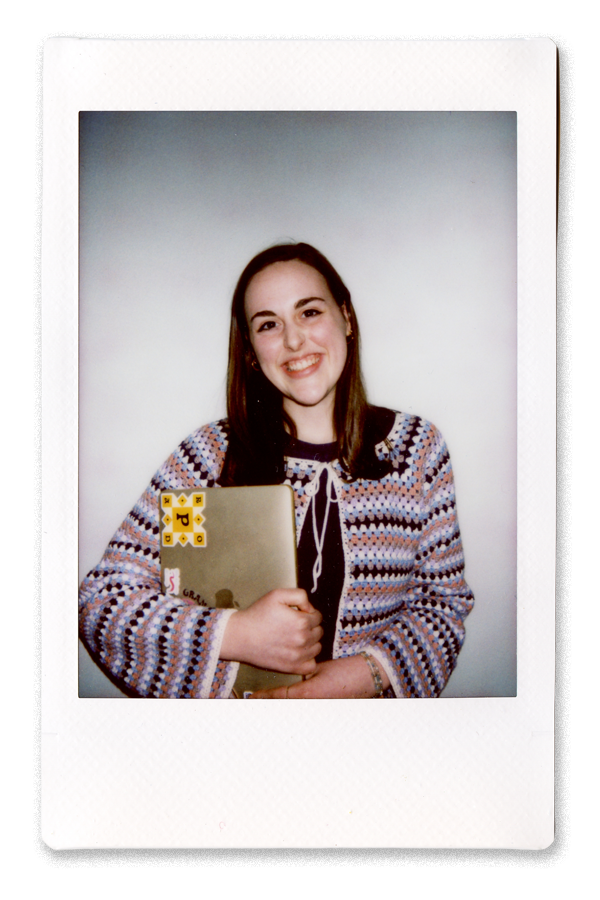 A young woman with long brown hair smiling and holding a laptop with stickers, standing against a white background.