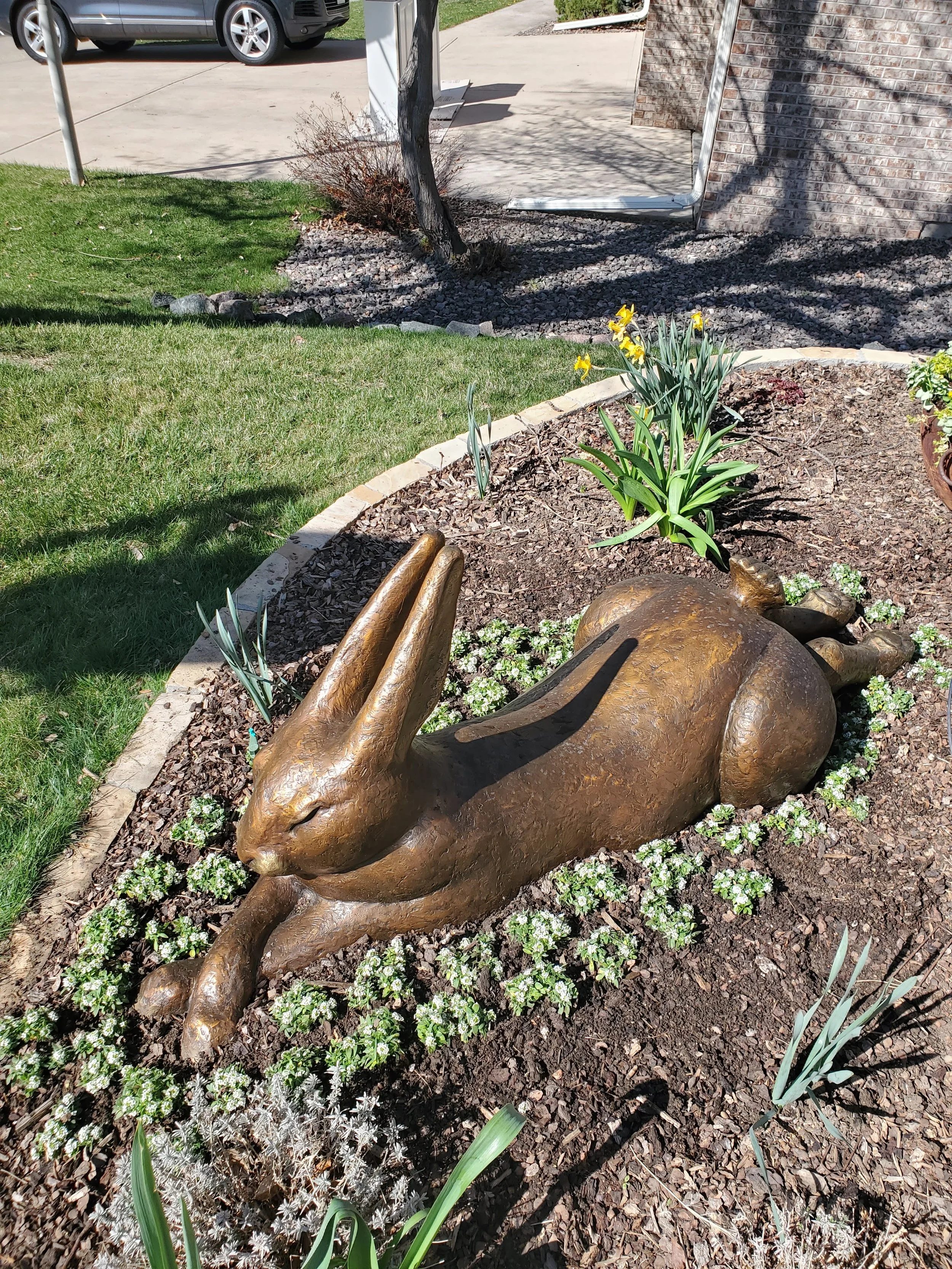 A bronze sculpture of a rabbit lying down in a garden bed, surrounded by plants and flowers.