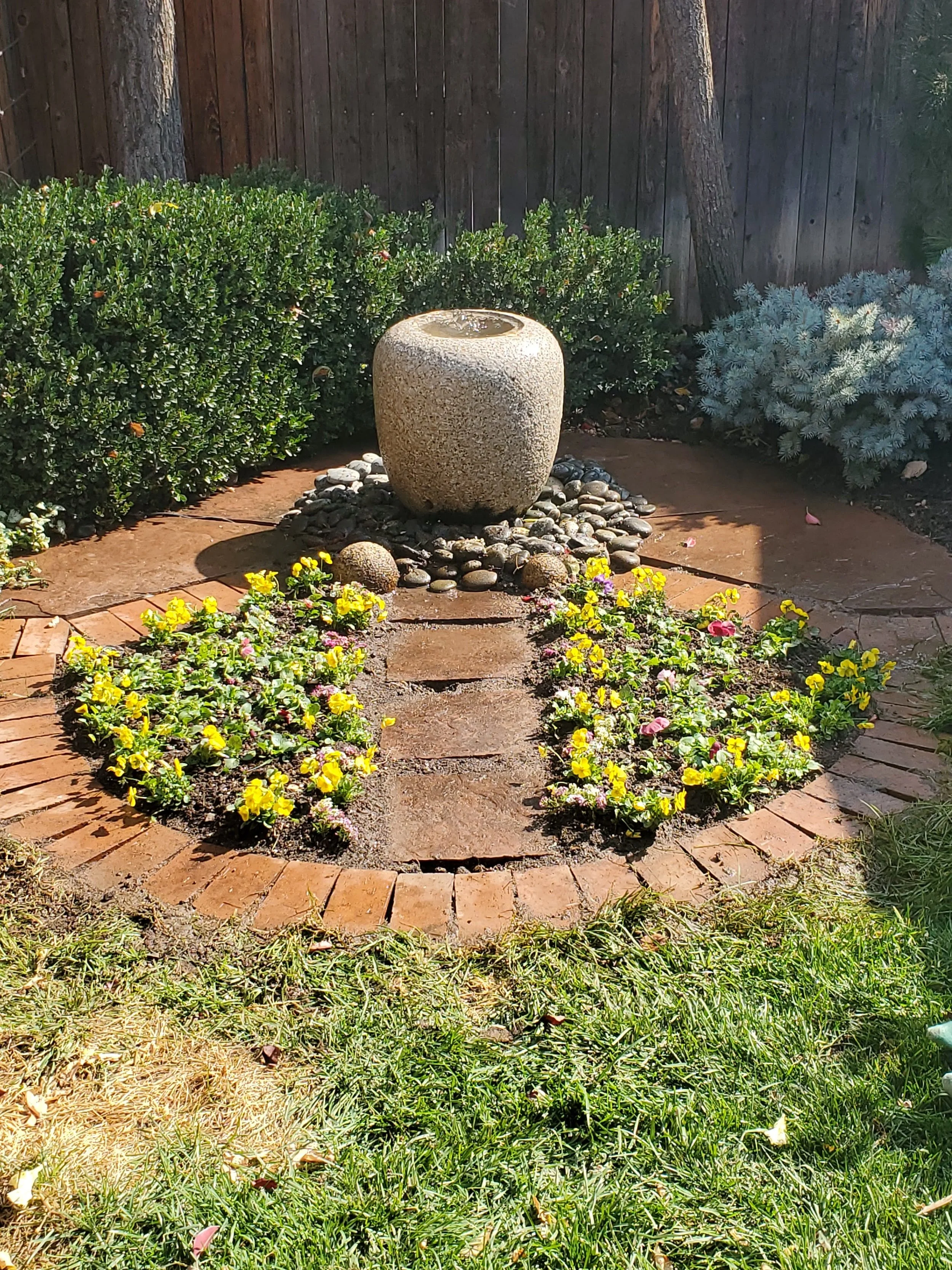 A garden with a stone water fountain, surrounded by yellow and pink flowers, bushes, and a wooden fence background.