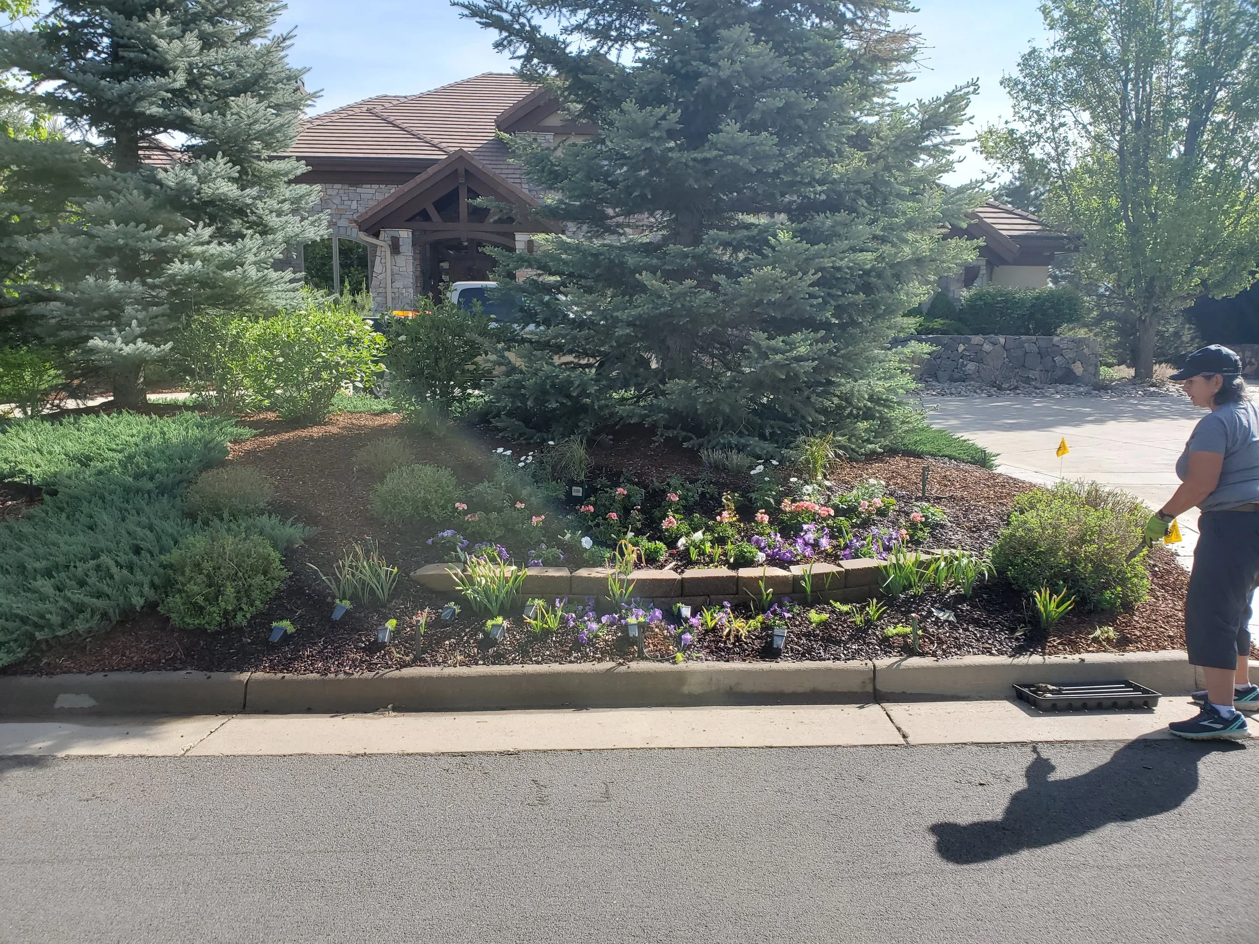 A person wearing a gray shirt, navy cap, and yellow gloves planting or tending to a garden bed with colorful flowers and shrubs in front of a house with stone walls and a sloped roof.