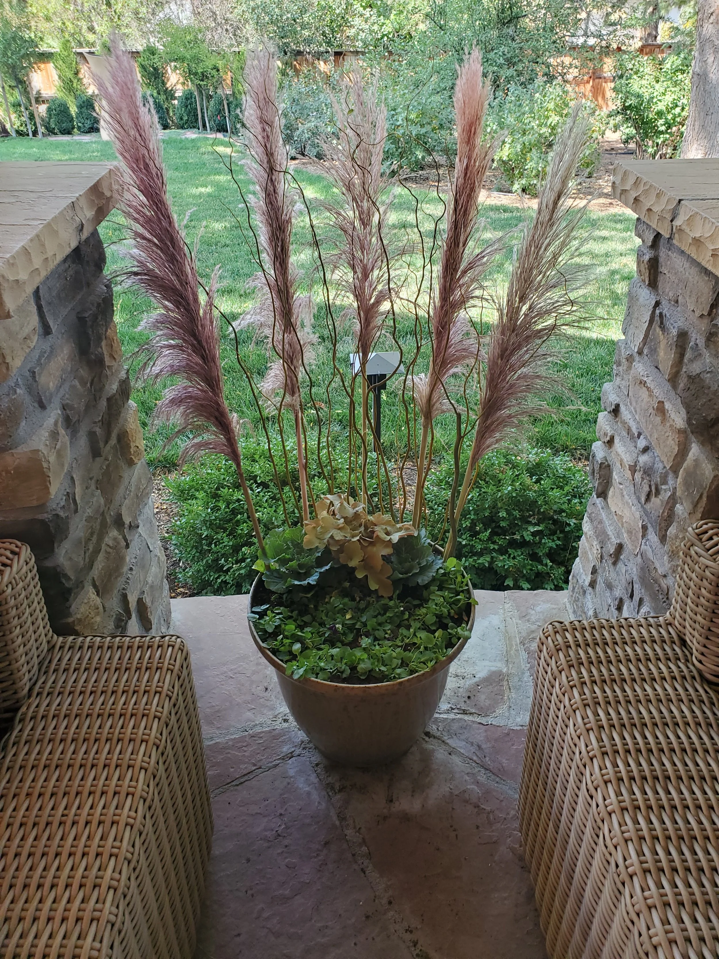A large potted plant with ornamental grasses and decorative foliage, flanked by two wicker chairs, on a stone porch with a garden and trees in the background.