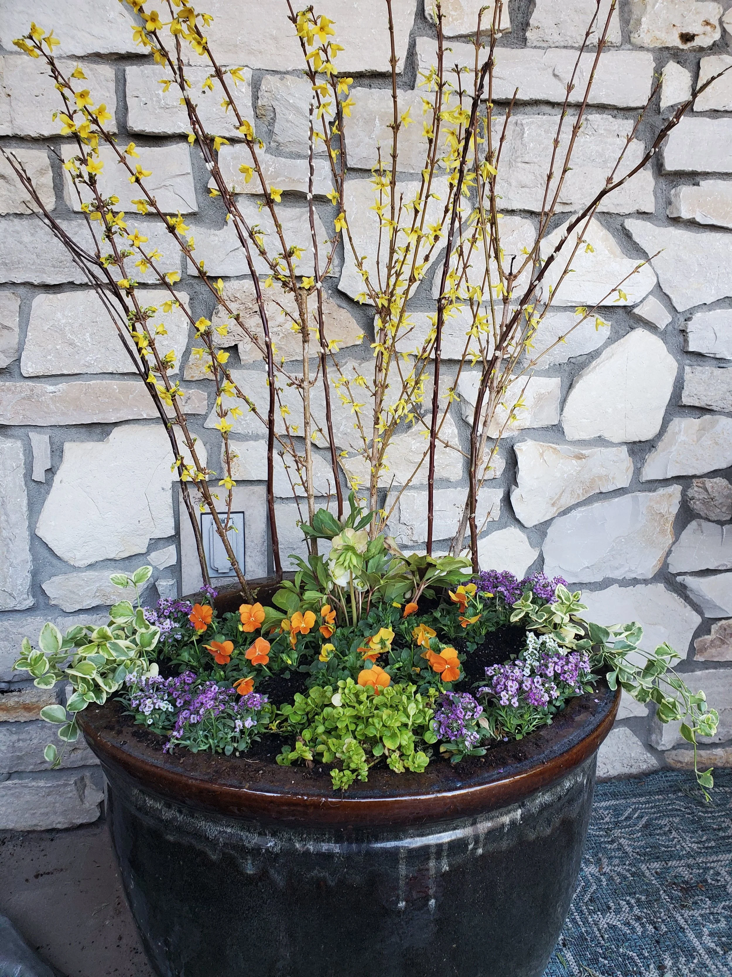 A large flower pot with yellow, orange, purple, and variegated green and white plants set against a stone wall.