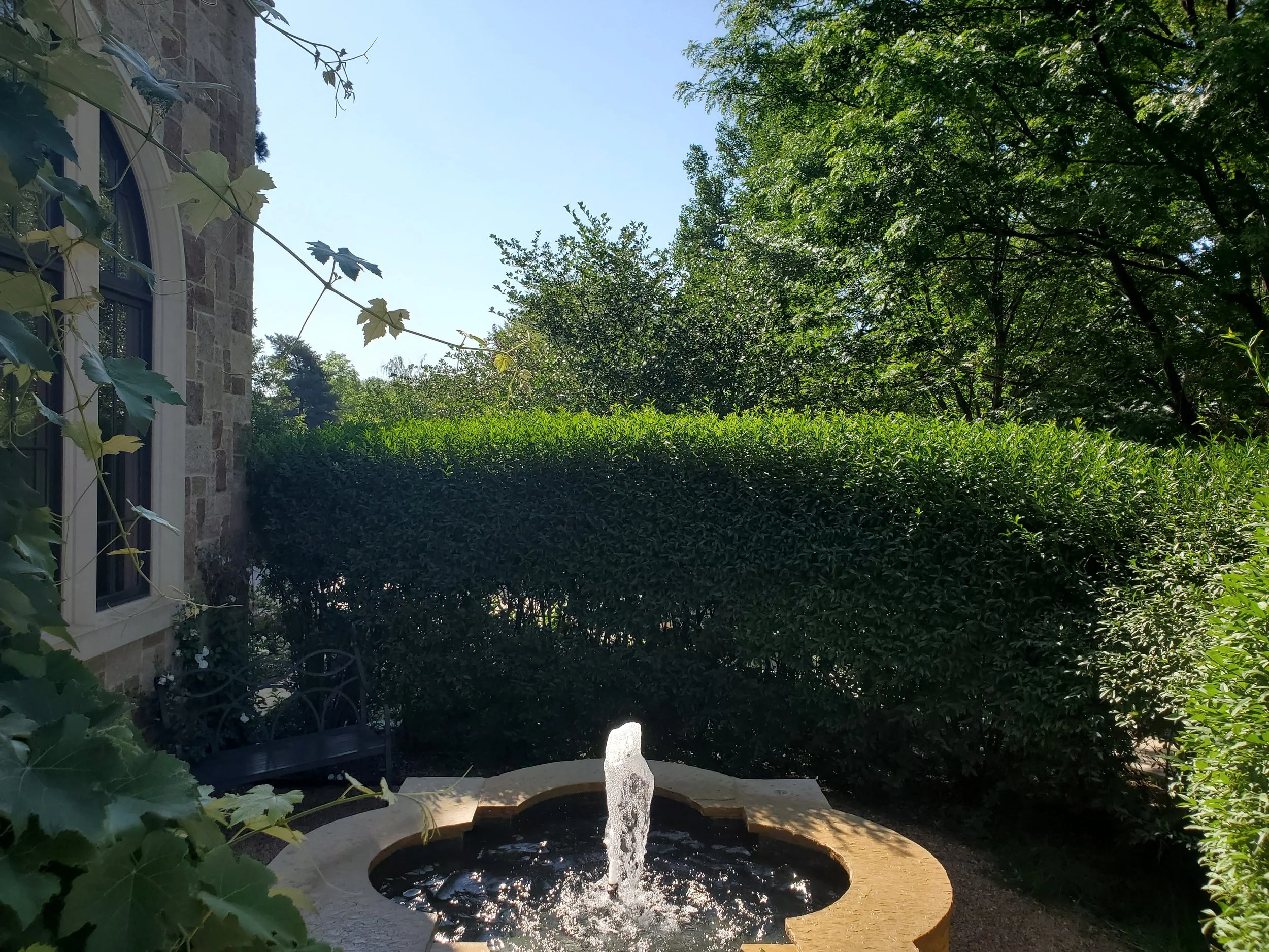 A small fountain with water bubbling up, in a garden area with bushes and trees, next to a stone house with a window and a black metal bench.