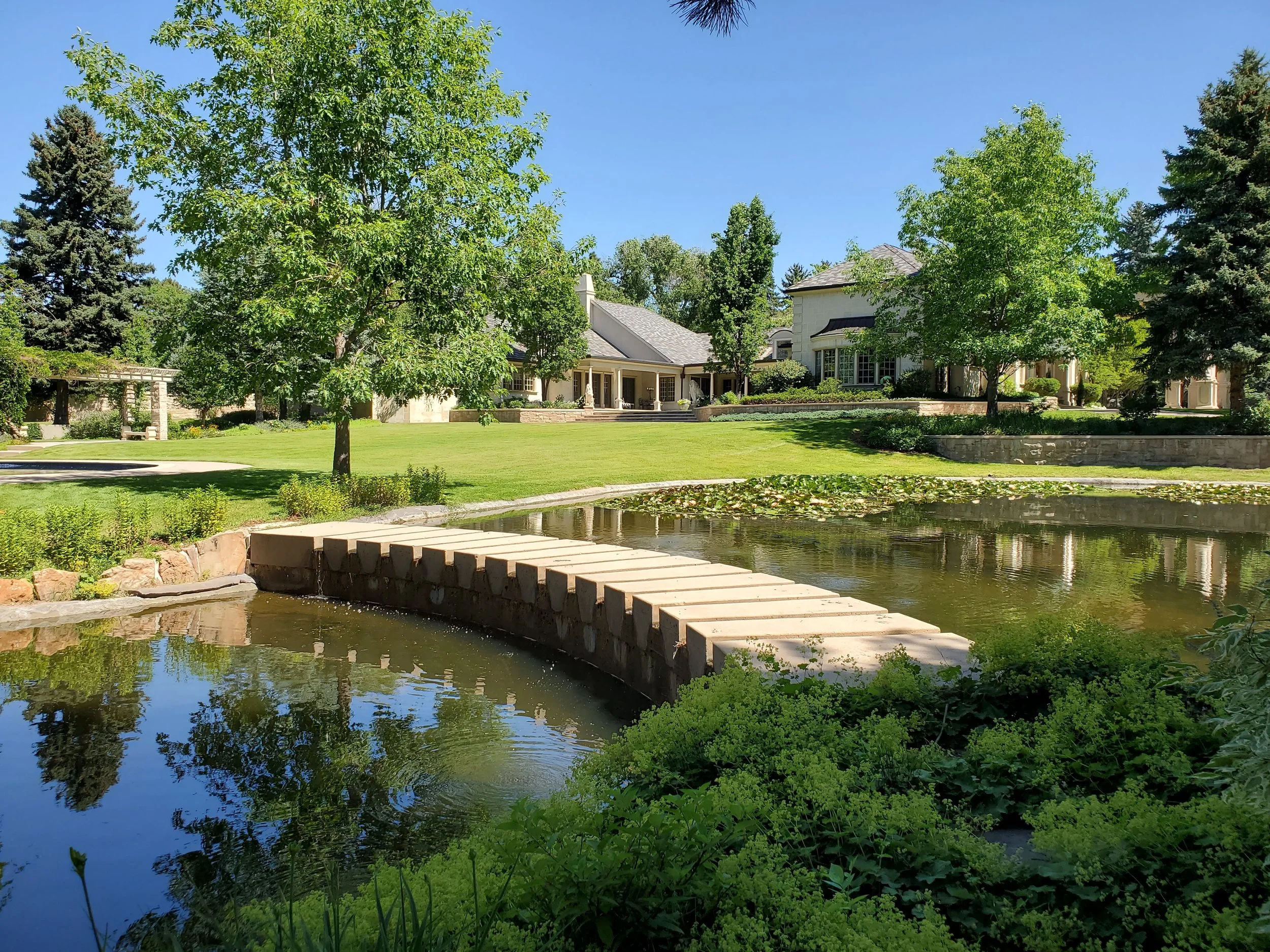 A large, luxury house with a well-kept lawn, trees, a pond with a small stone bridge, and landscaping on a sunny day.