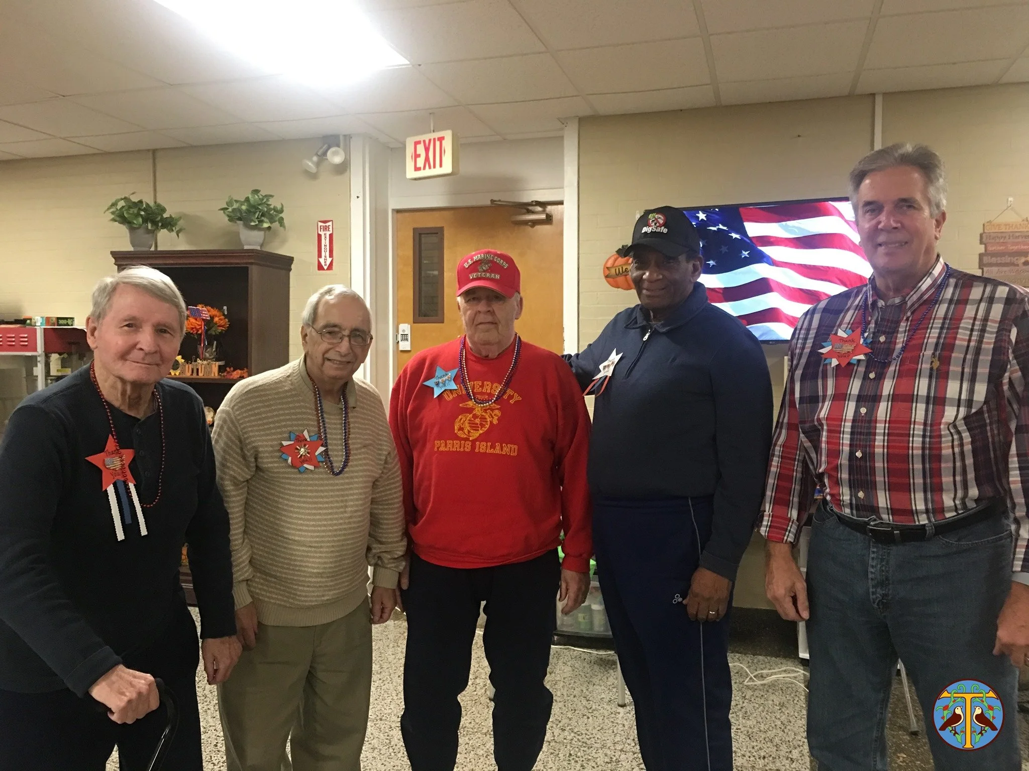Five men standing indoors, dressed casually, wearing patriotic decorations, with an American flag on a screen behind them.