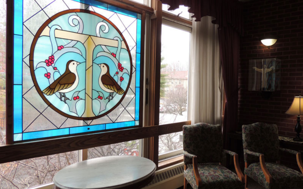 Stained glass window depicting two birds perched on branches with red berries, inside a room with chairs, curtains, and a brick wall.