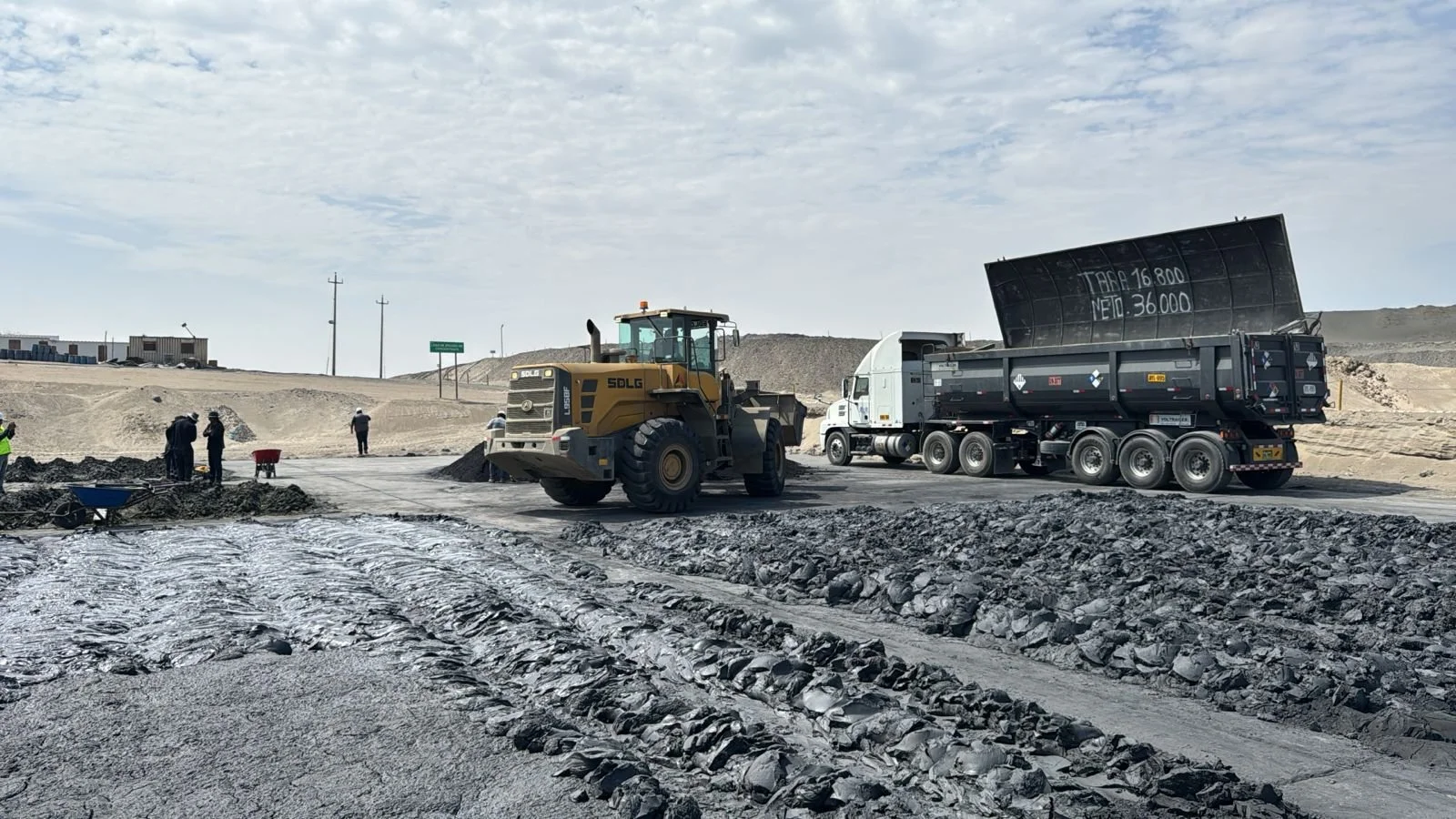 Construction site with dump truck unloading materials, bulldozer, and workers on a dirt road with a cloudy sky.