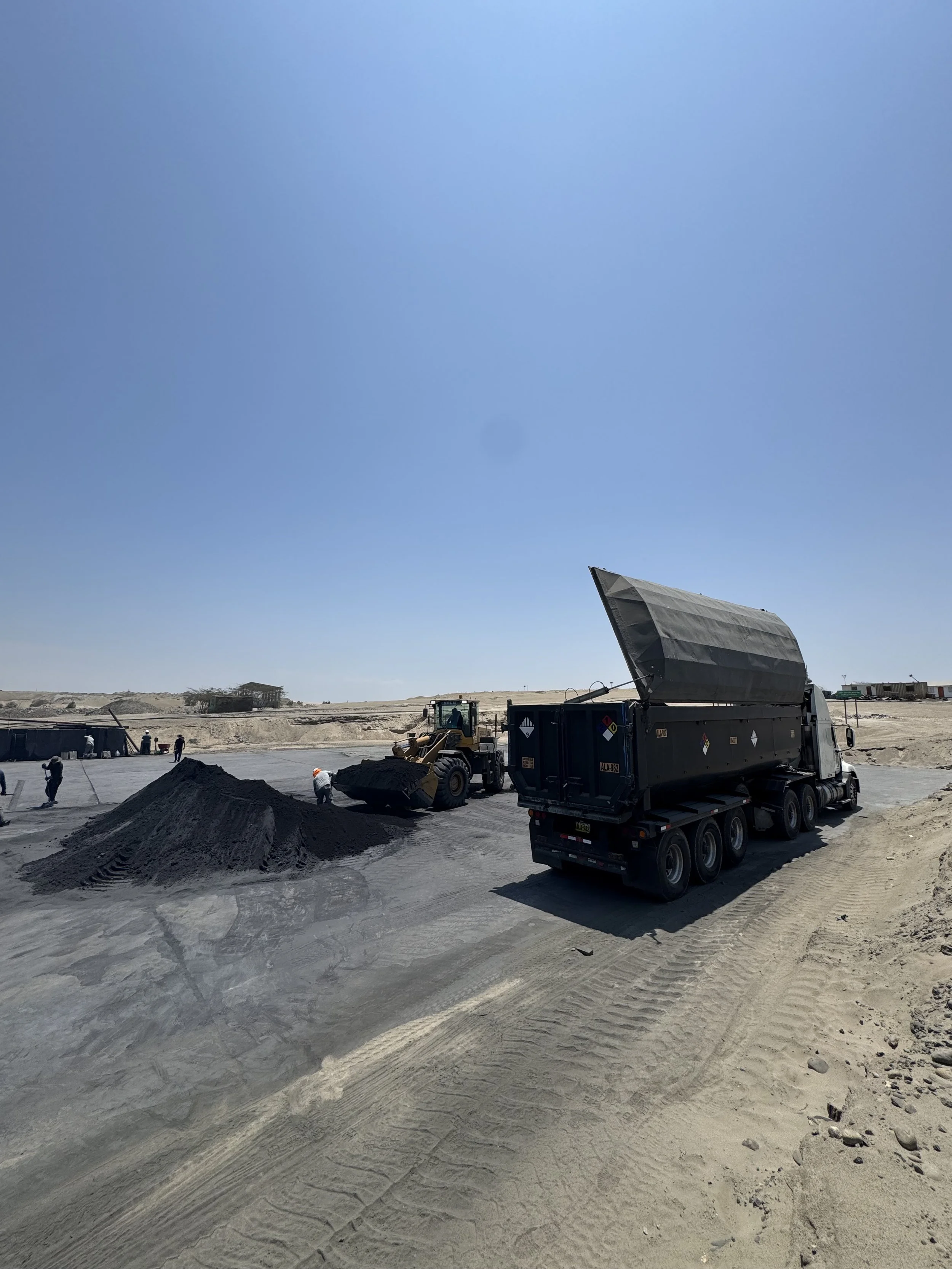Construction site in a desert with a large truck and a bulldozer working on asphalt, and several workers nearby.