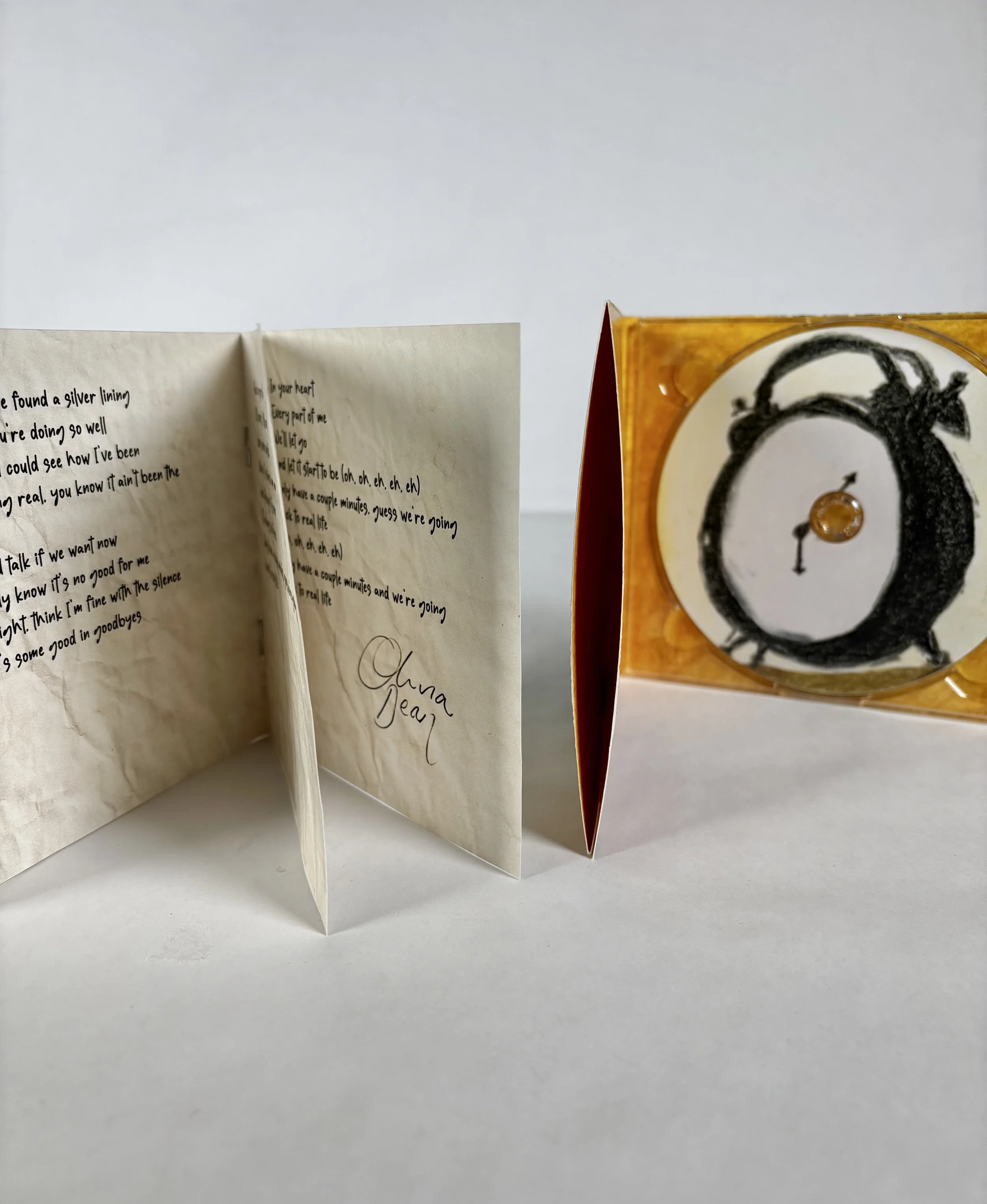 Open handwritten note on cream-colored paper and a small square-shaped container with black drawing of a clock with an orange-colored circular element inside.