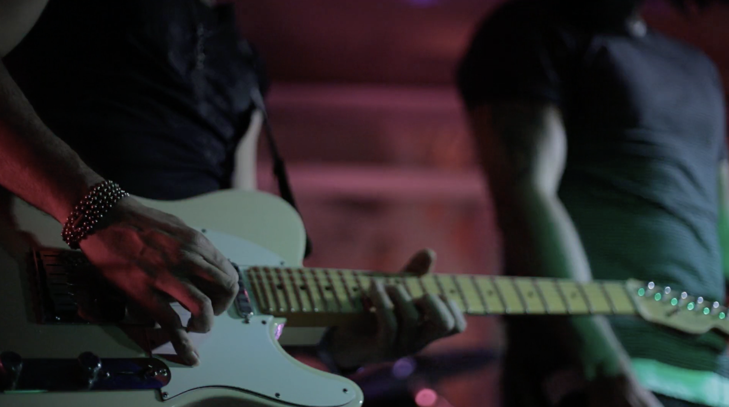 Close-up of a guitarist's hand playing an electric guitar during a live performance, with another musician in the background.