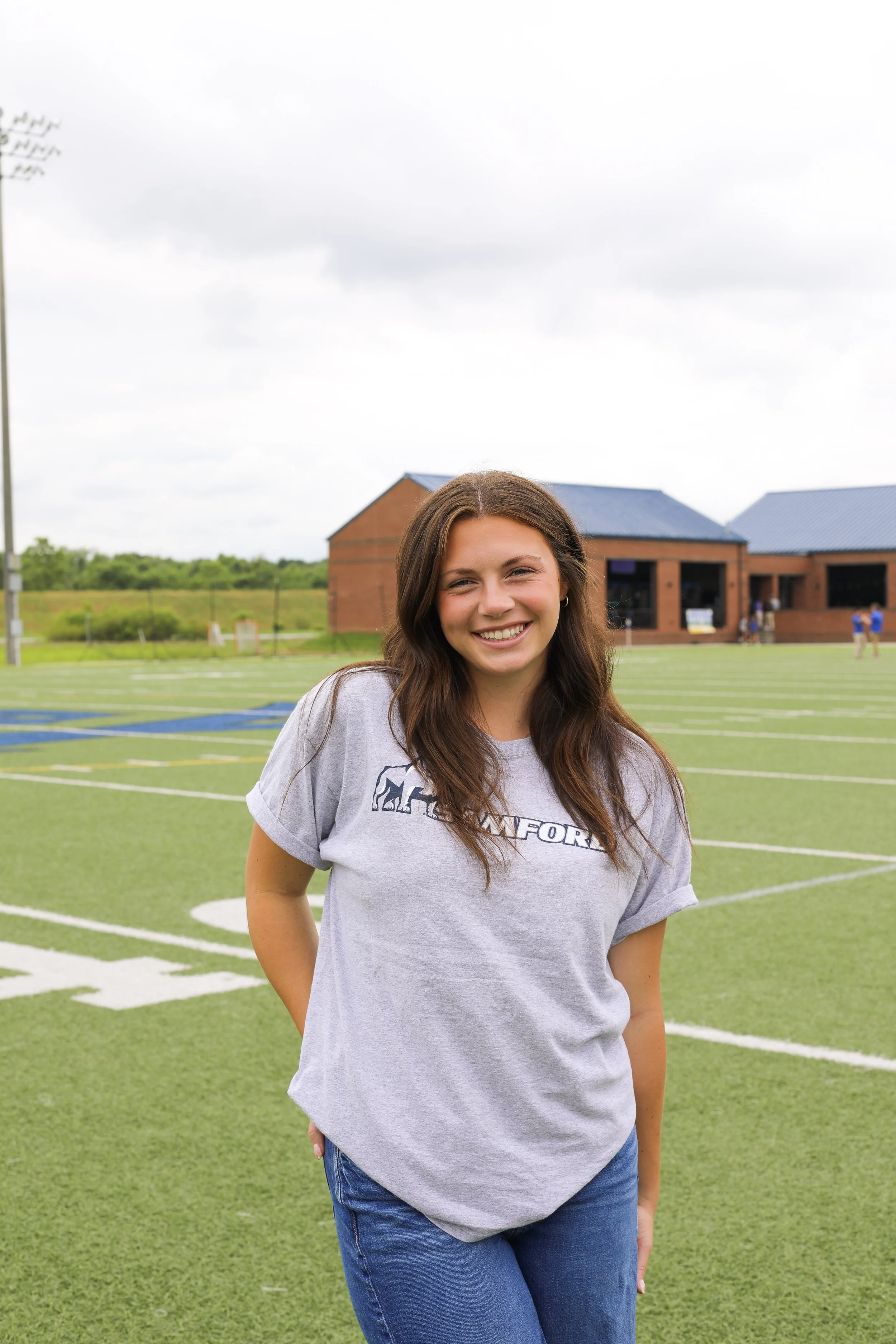 Young woman standing on a football field, smiling, with a school building and cloudy sky in the background.