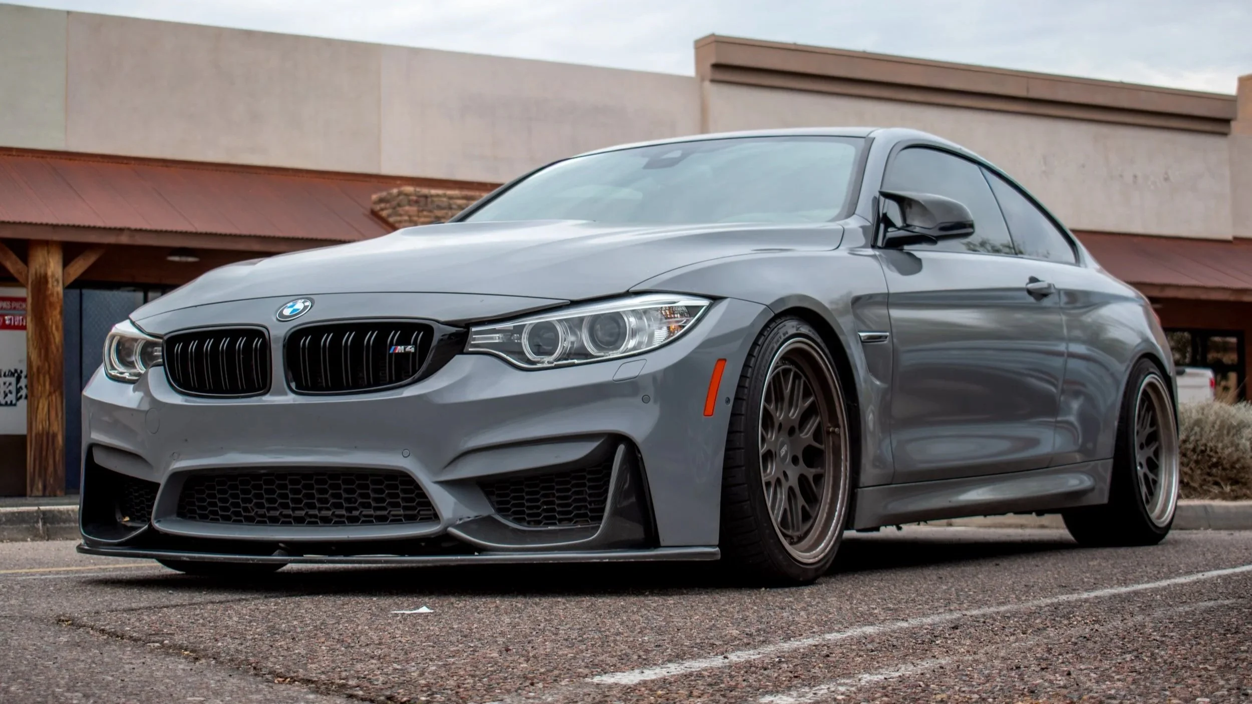 Gray BMW M4 sports car parked in a lot with a building in the background.