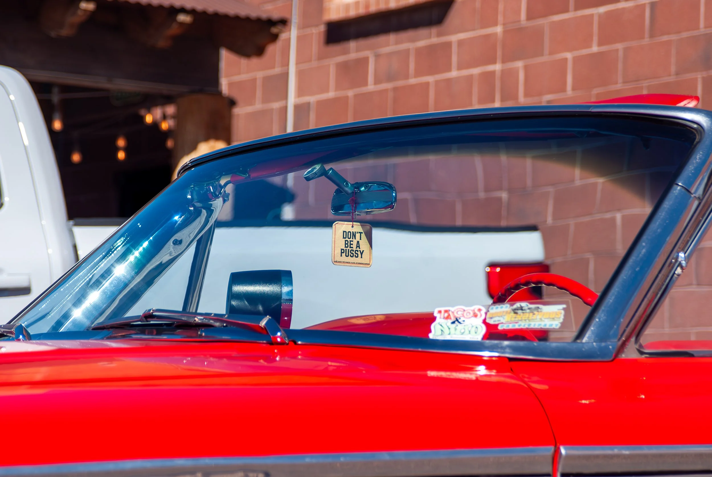 Close-up of a vintage red car with a hanging air freshener that says 'Don't Be A Pussy' inside, with stickers on the dashboard visible through the windshield.