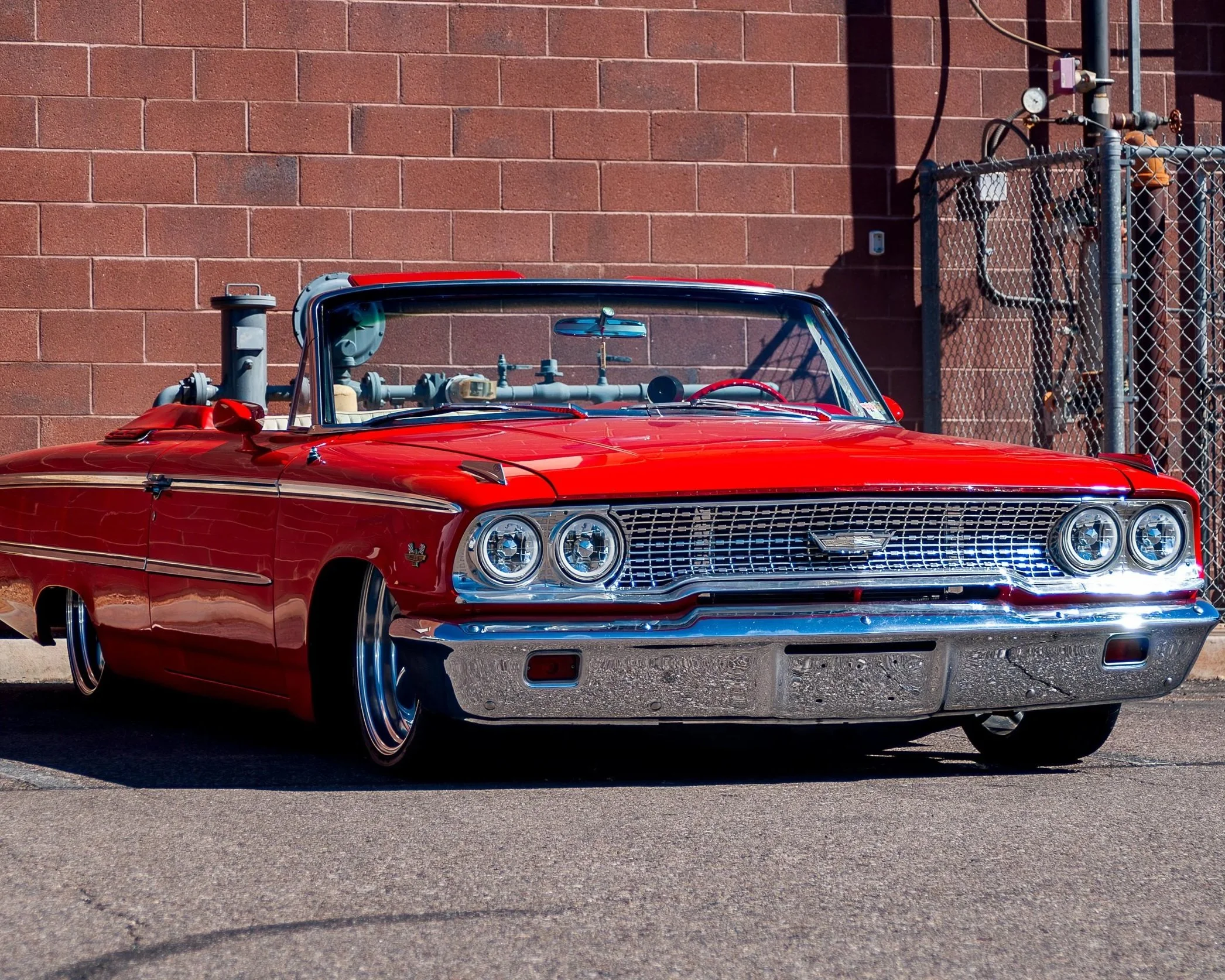 Red vintage convertible car parked on the street near a brick wall and chain-link fence, with visible engine parts and chrome accents.