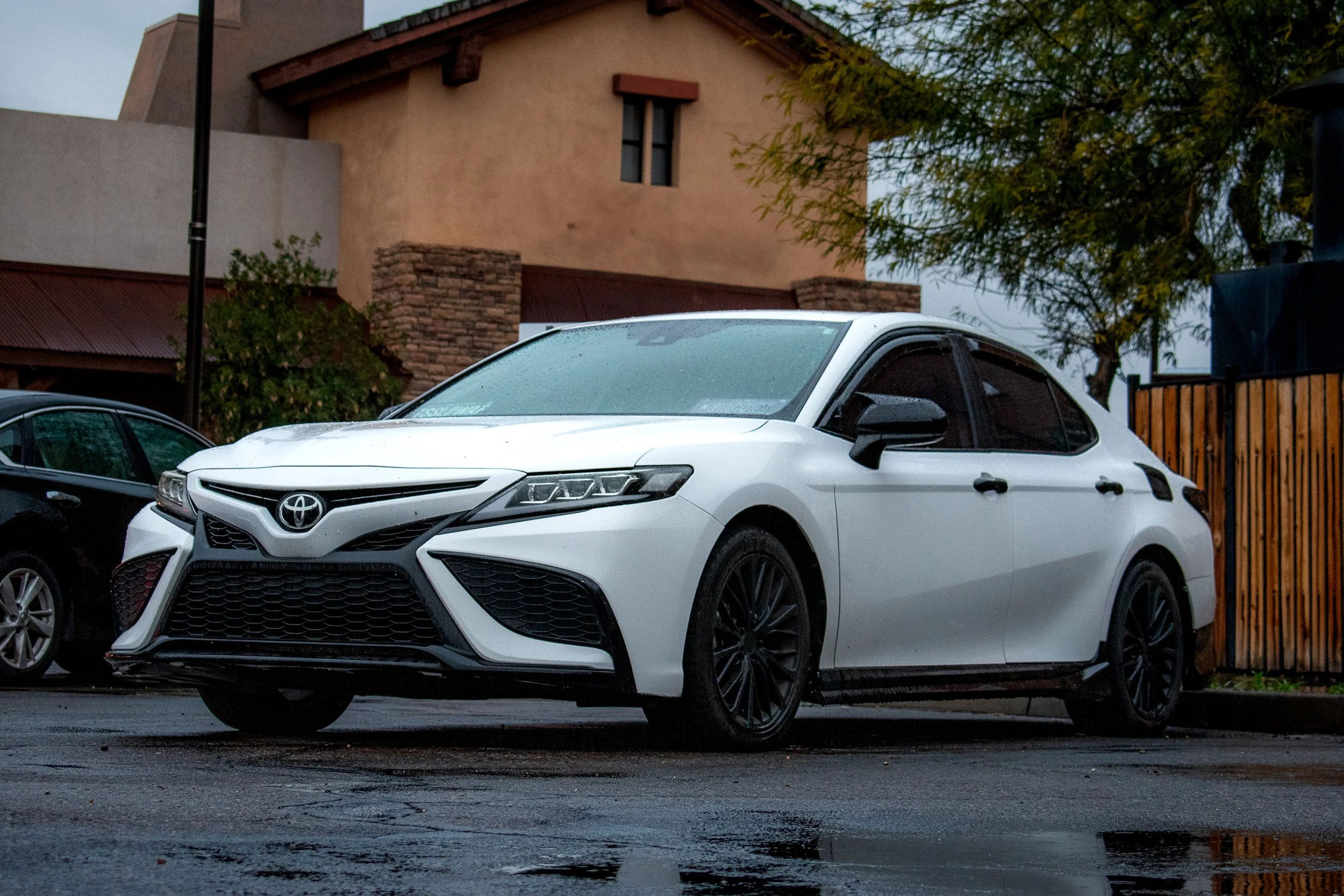 A white Toyota sedan parked on a wet street near a house with a tan stucco exterior and stone accents, during rainy weather.