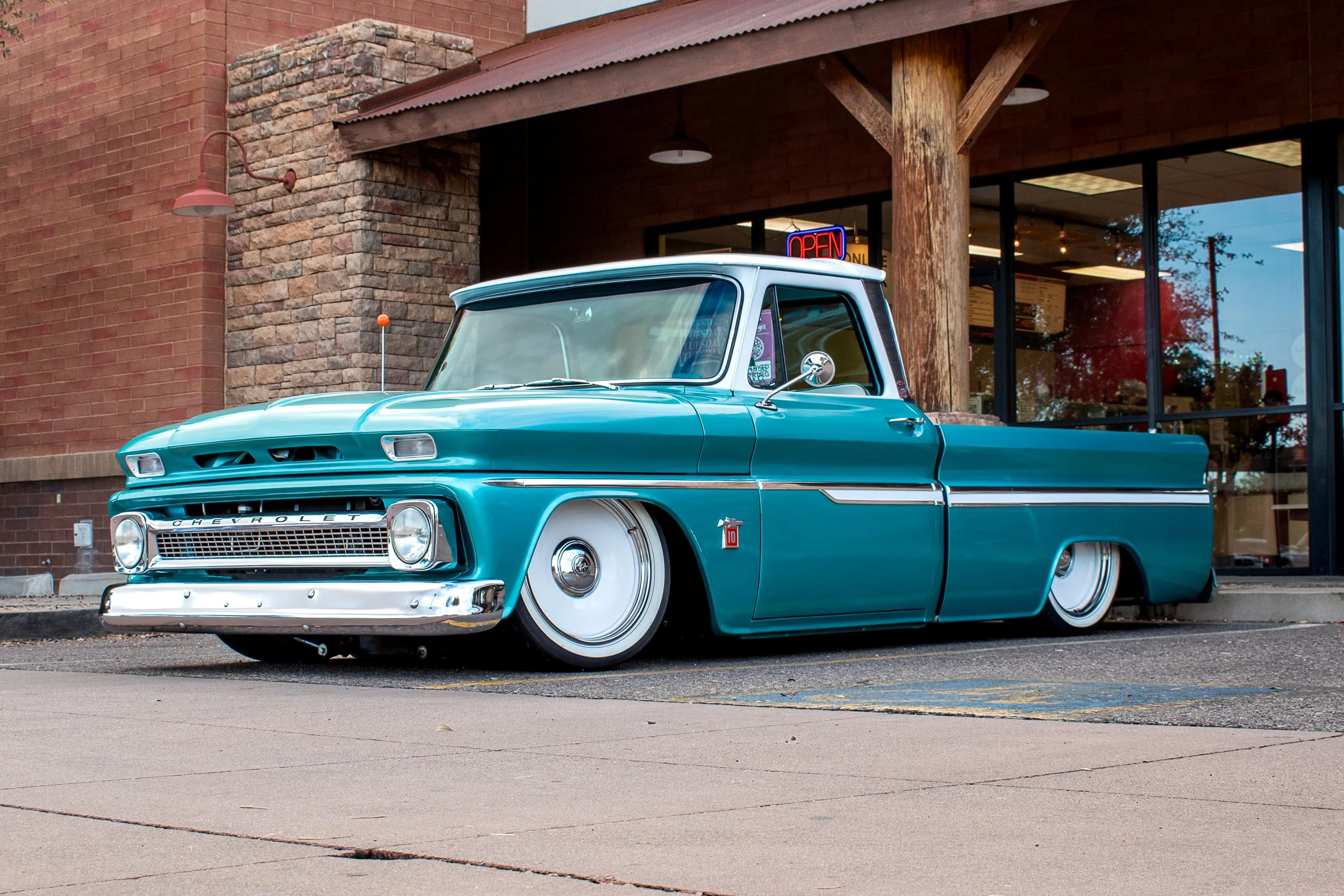 A vintage Chevrolet pickup truck painted in teal parked in front of a storefront with a neon 'Open' sign.