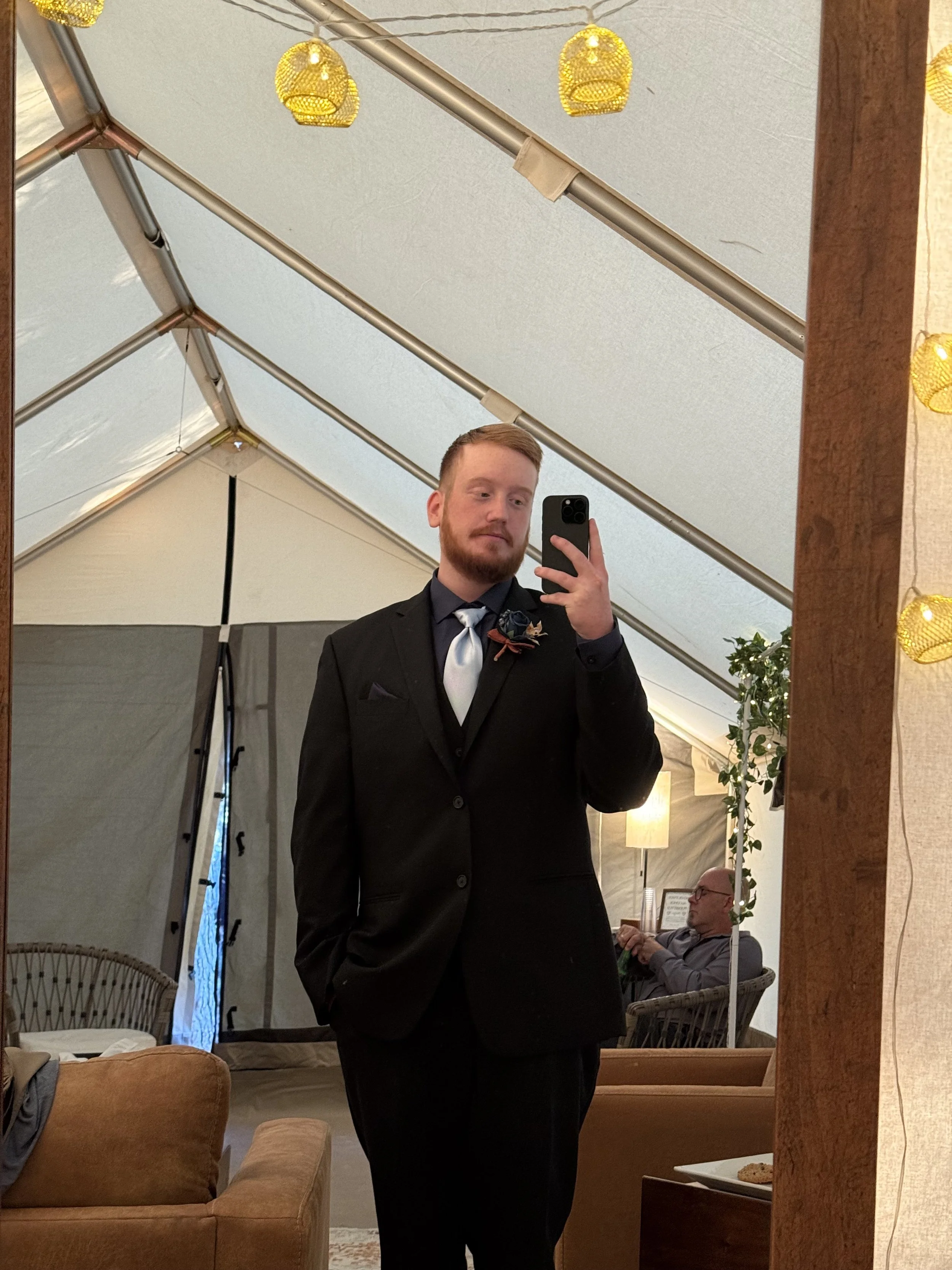 A man dressed in a black suit with a light blue tie taking a selfie in a large mirror at an indoor event tent.