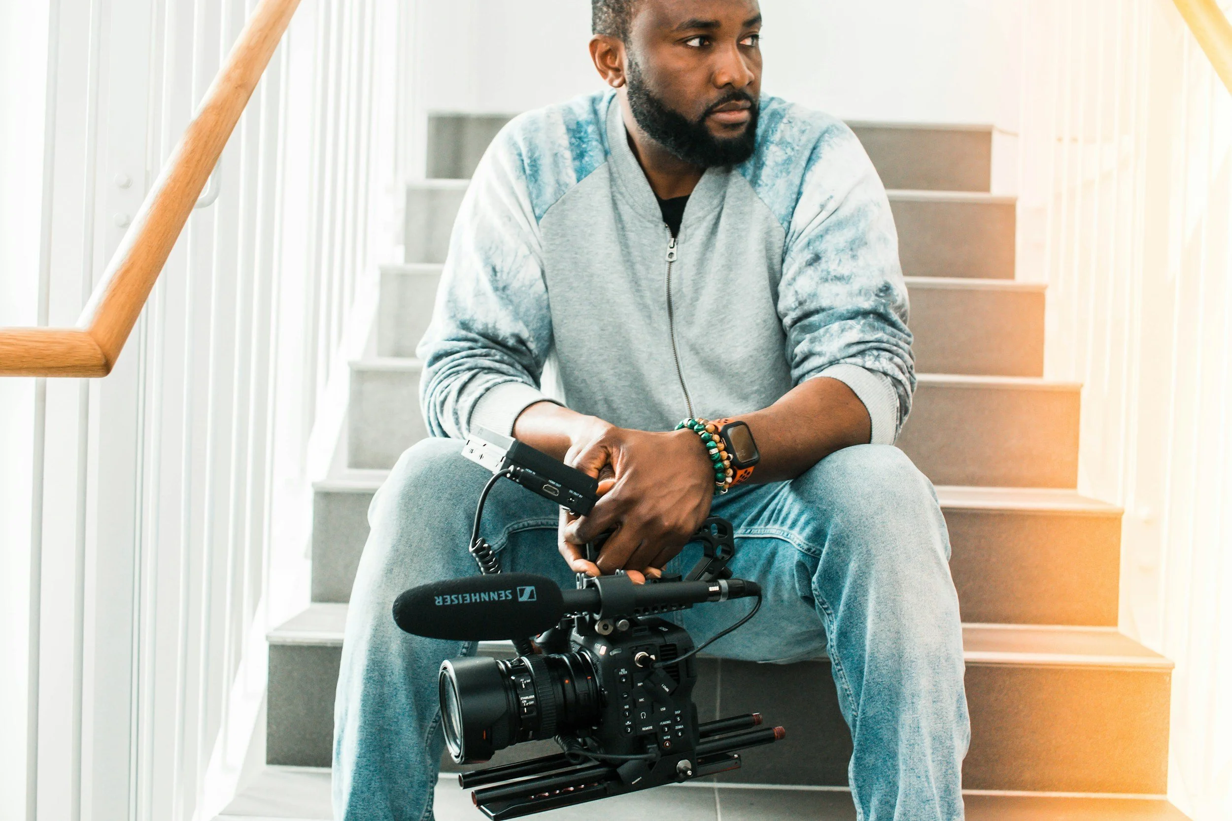 A man sitting on a stairway in Dubai holding a professional video camera with a microphone. He is wearing a gray sweatshirt, jeans, and a smartwatch, with a focused expression.