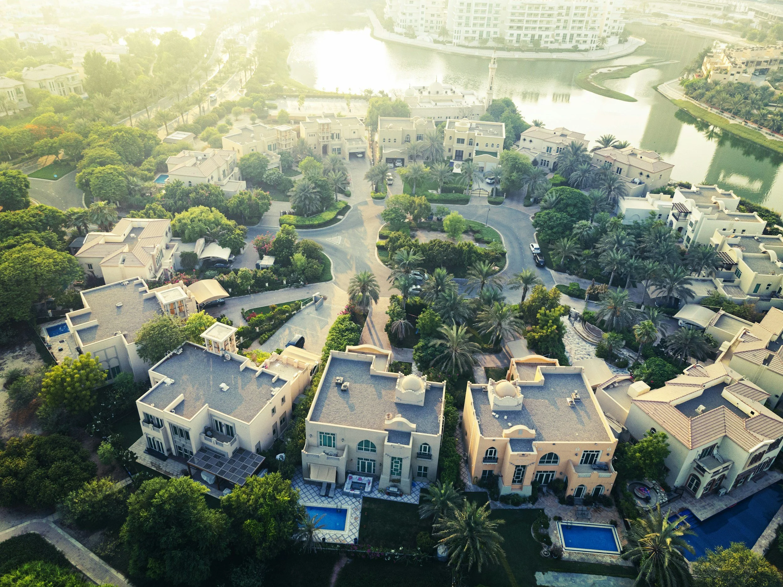 An aerial view of a luxury Dubai residential neighborhood with large houses, lush greenery, palm trees, swimming pools, and a body of water in the background.