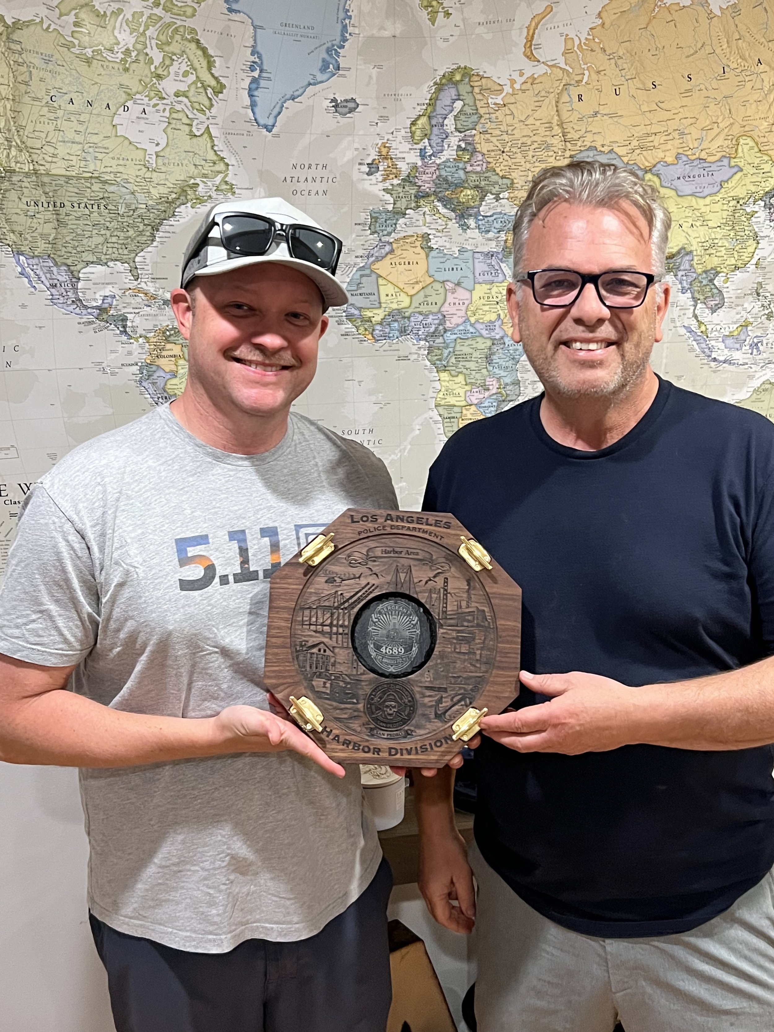 Two men smiling and holding a wooden plaque with a police badge engraving, standing in front of a world map. One man is wearing a gray T-shirt and a white cap with sunglasses, the other is in a black T-shirt and glasses.