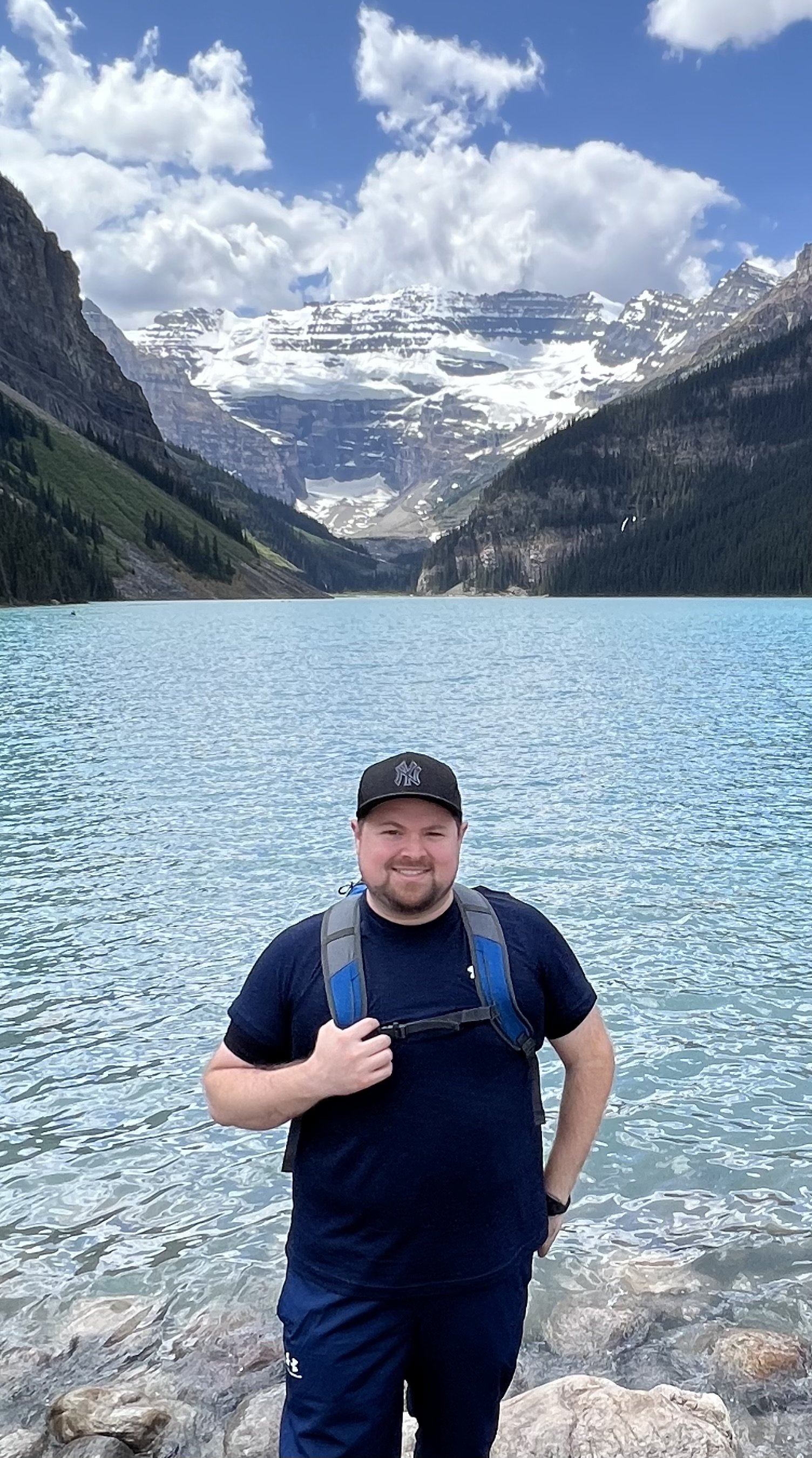 A man standing on rocks by a body of water with snow-capped mountains and a cloudy sky in the background.