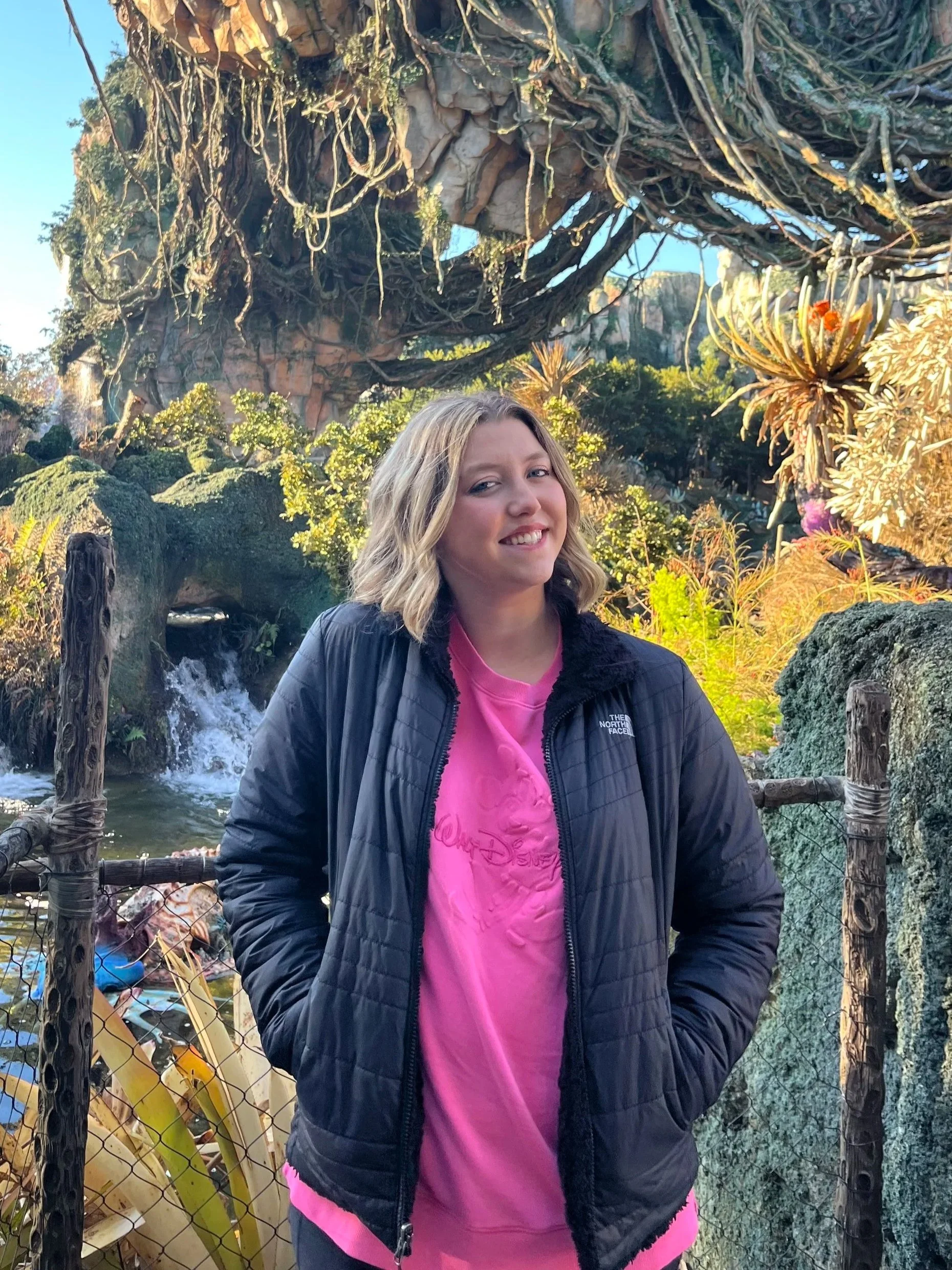A young woman with shoulder-length blonde hair smiling outdoors in front of a lush, themed landscape with waterfalls and large artificial floating mountain in the background, wearing a black North Face jacket over a pink Disney t-shirt.