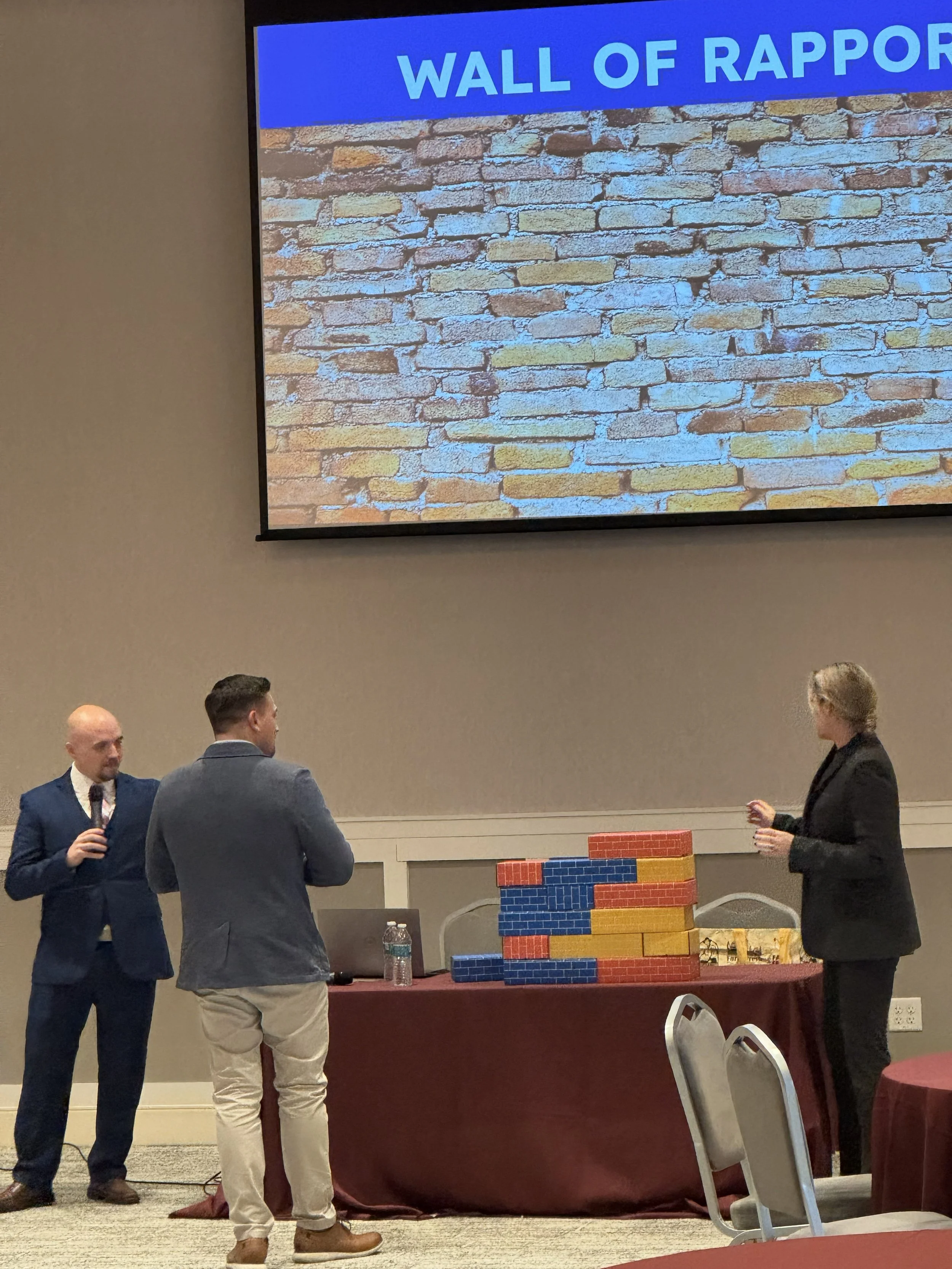 Three people standing in front of a table with large colored bricks in a conference room. One person is speaking into a microphone, and a presentation slide titled 'Wall of Rapport' is projected on the wall behind them.
