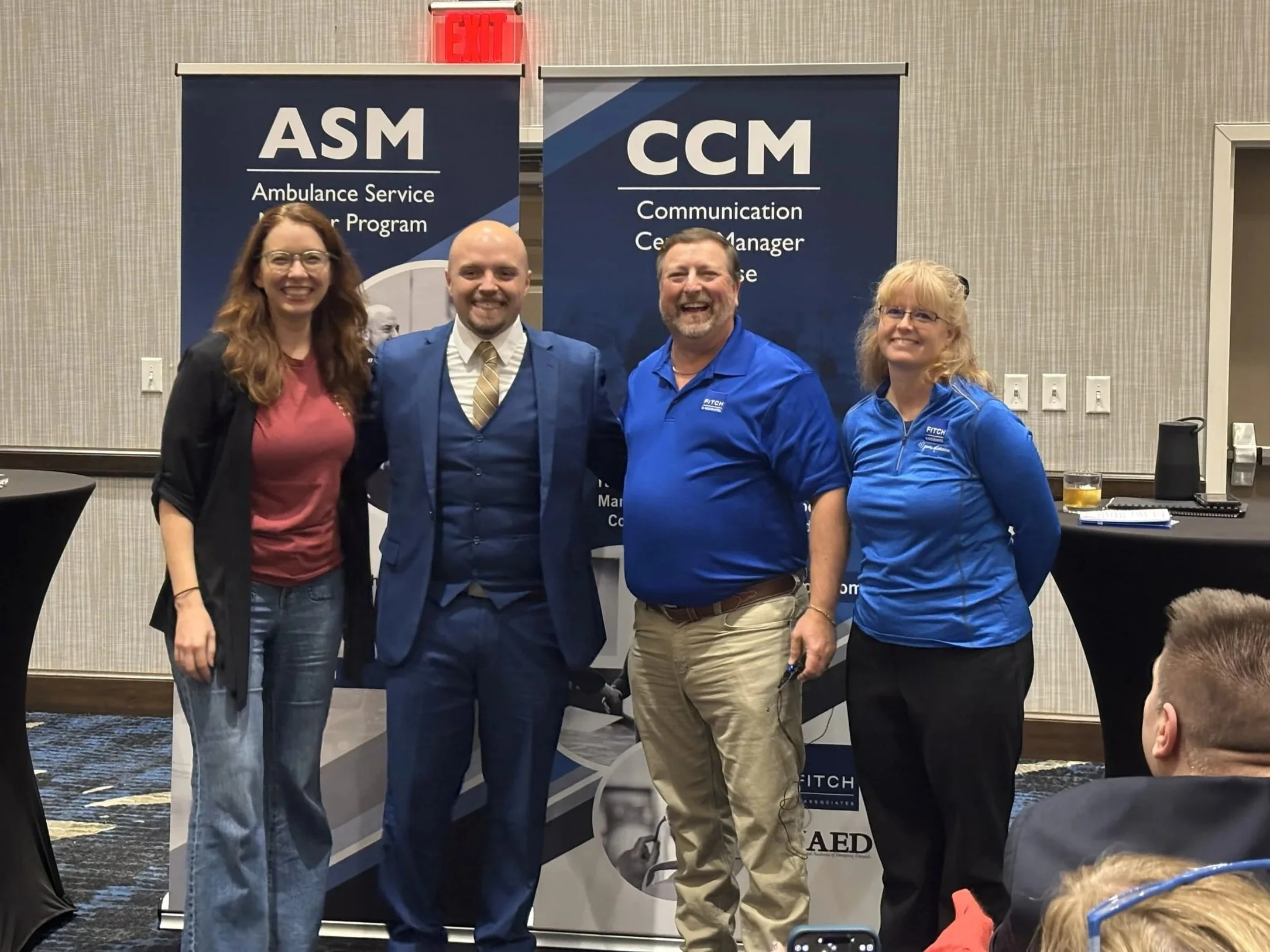 Four people standing together at a professional event, smiling in front of two blue banners. The left banner reads 'ASM Ambulance Service' and the right banner reads 'CCM Communication Center Manager.' The group includes two women and two men, dressed in business casual and business attire.