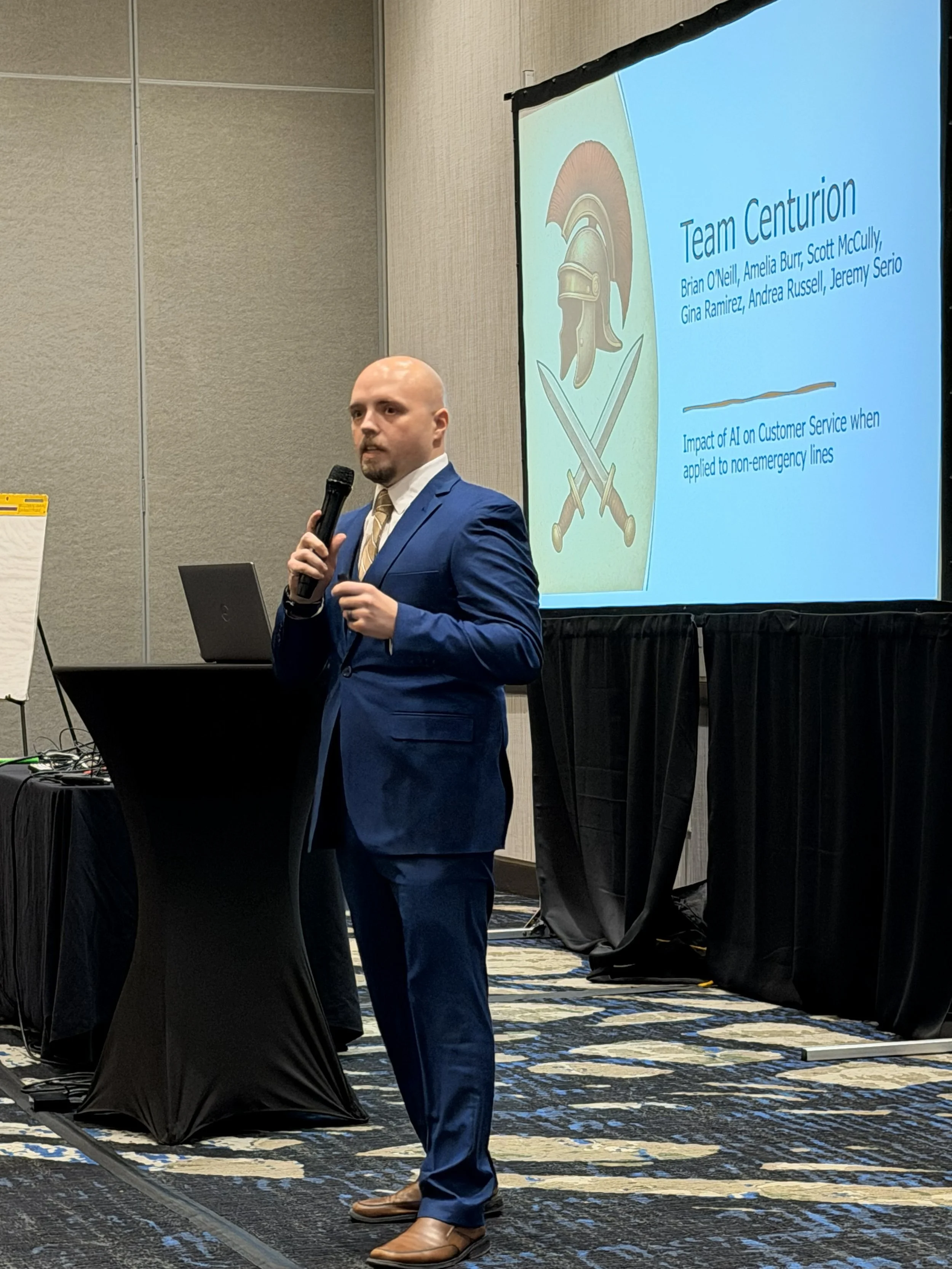 A man in a blue suit holding a microphone standing next to a presentation screen during a conference.